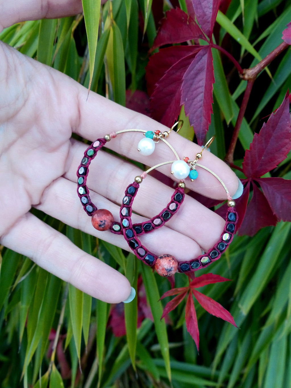 Peut inclure: Une paire de grandes boucles d'oreilles créoles avec un cordon bordeaux et des perles noires et irisées. Les boucles d'oreilles ont un anneau en or et un charme suspendu avec une perle blanche et des perles turquoise et rouges.