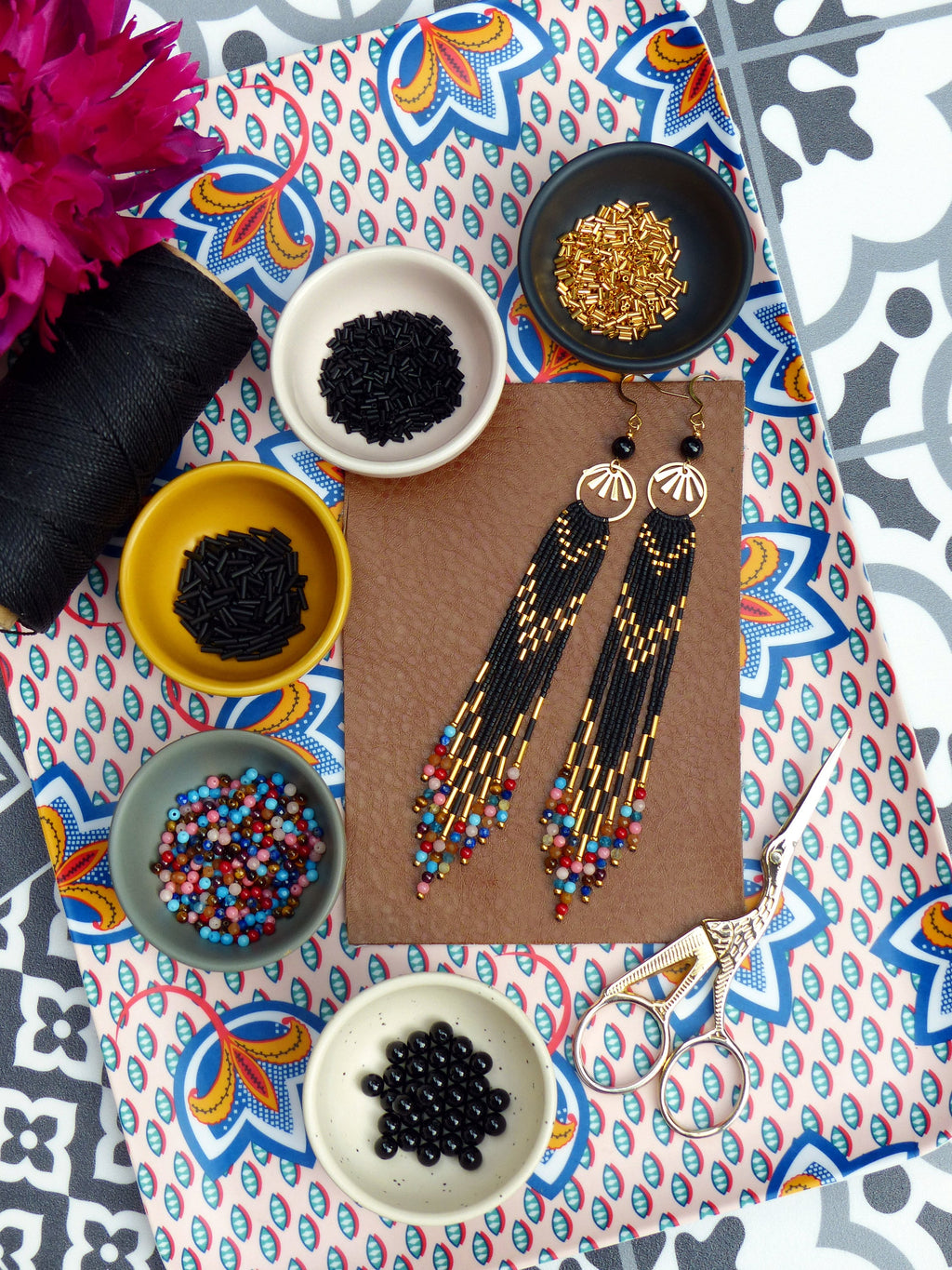 Peut inclure: Un plateau avec divers bols de perles, y compris des perles noires, dorées et multicolores. Il y a aussi deux paires de longues boucles d'oreilles noires perlées avec des accents dorés. Le plateau a un motif floral coloré.
