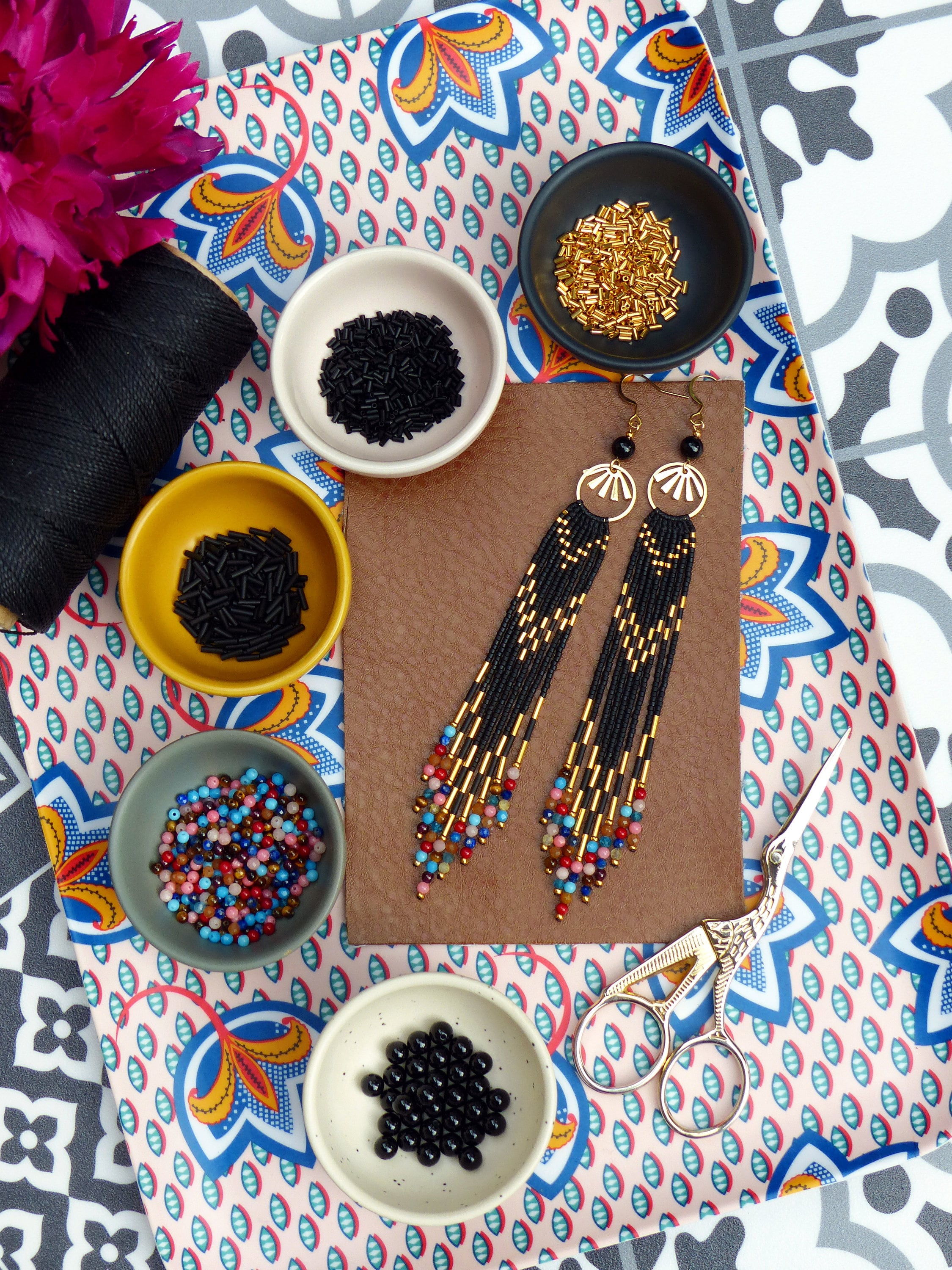 Peut inclure: Un plateau avec divers bols de perles, y compris des perles noires, dorées et multicolores. Il y a aussi deux paires de longues boucles d'oreilles noires perlées avec des accents dorés. Le plateau a un motif floral coloré.