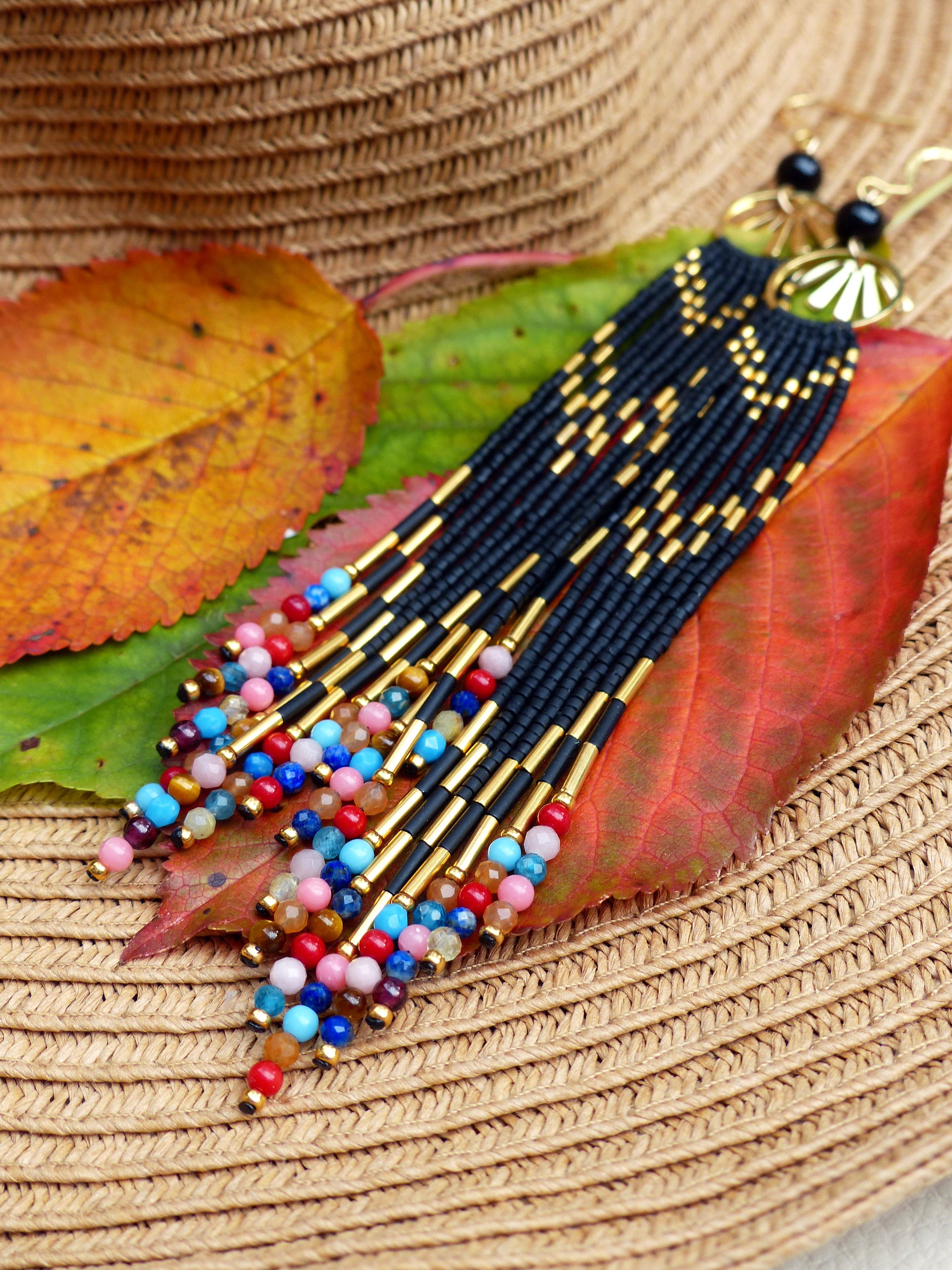 Peut inclure: Longues boucles d'oreilles noires en perles avec des accents dorés et des perles de pierres précieuses colorées. Les boucles d'oreilles sont présentées sur un chapeau de paille et tombent sur des feuilles d'automne.
