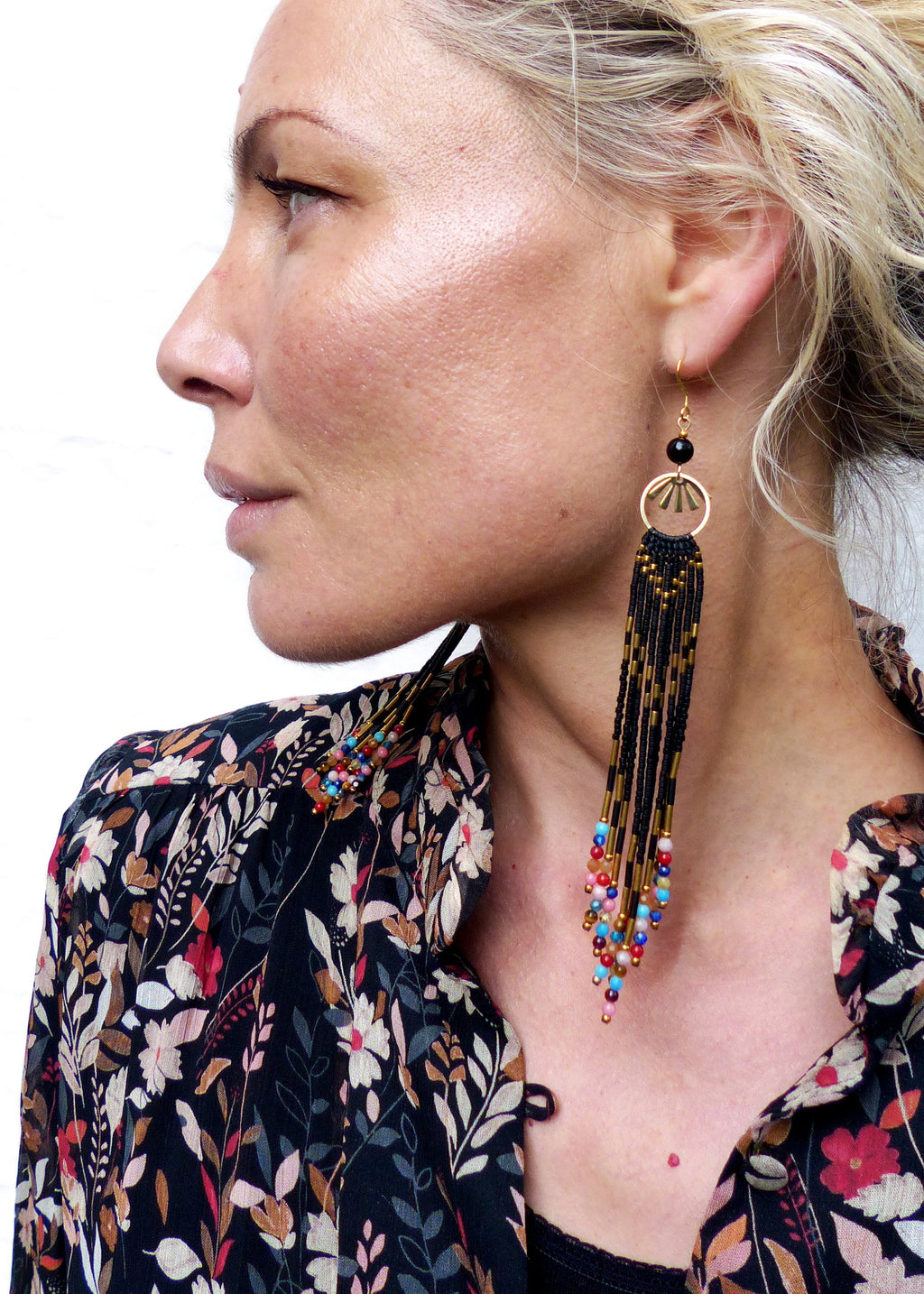 Peut inclure: Longues boucles d'oreilles noires en perles avec des accents dorés et des perles colorées qui pendent du bas. Les boucles d'oreilles sont portées par une personne aux cheveux blonds.