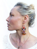 Peut inclure: Une femme portant un haut en crochet blanc et un collier en or avec un pendentif en forme de croix en cristal. Elle porte de longues boucles d'oreilles en perles avec un motif géométrique en rouge, orange, jaune, bleu et noir.