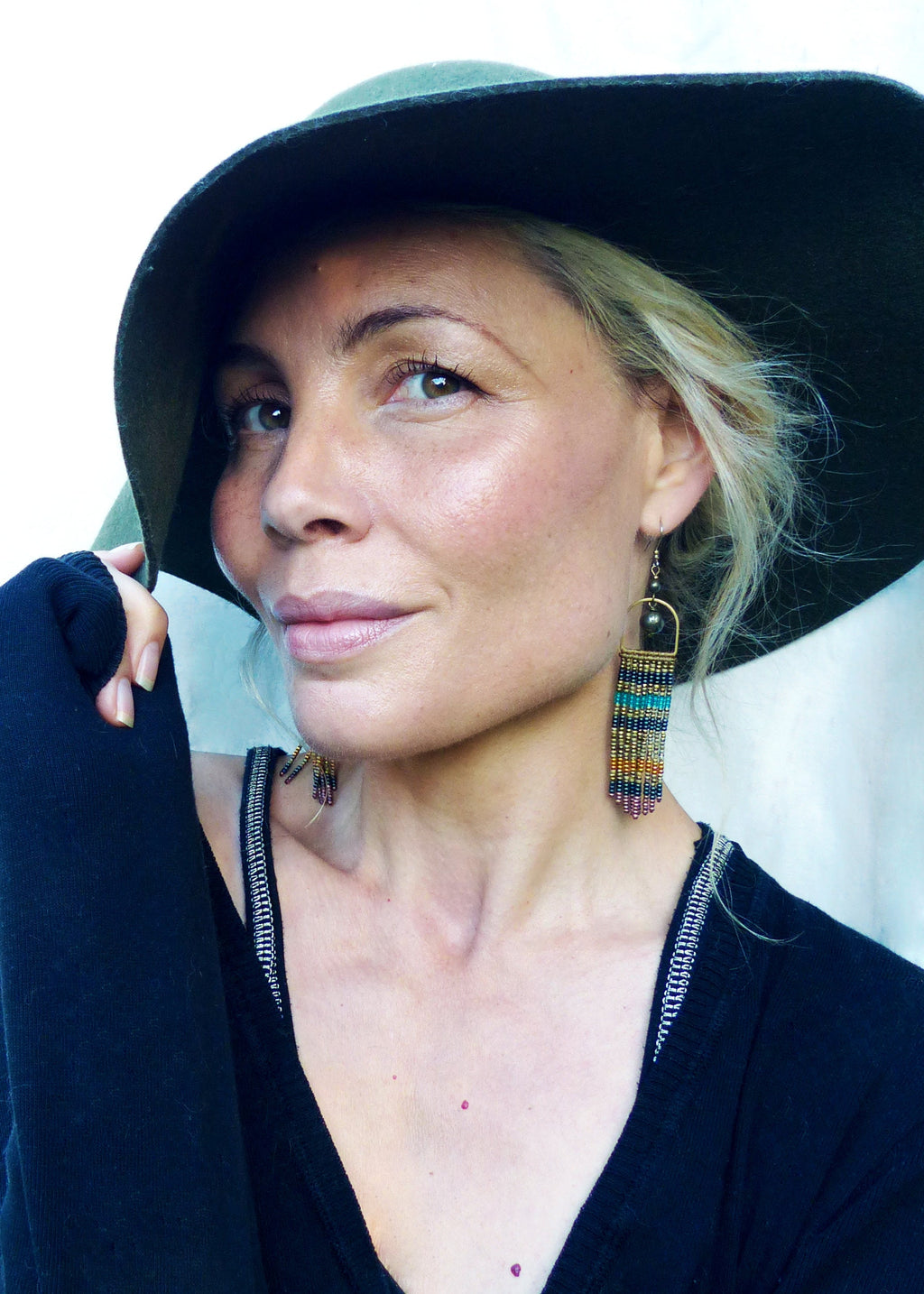 Peut inclure: Une femme portant un chapeau en feutre vert et un pull noir avec un détail de fermeture éclair argentée. Elle porte de longues boucles d'oreilles en perles de couleur turquoise et or.