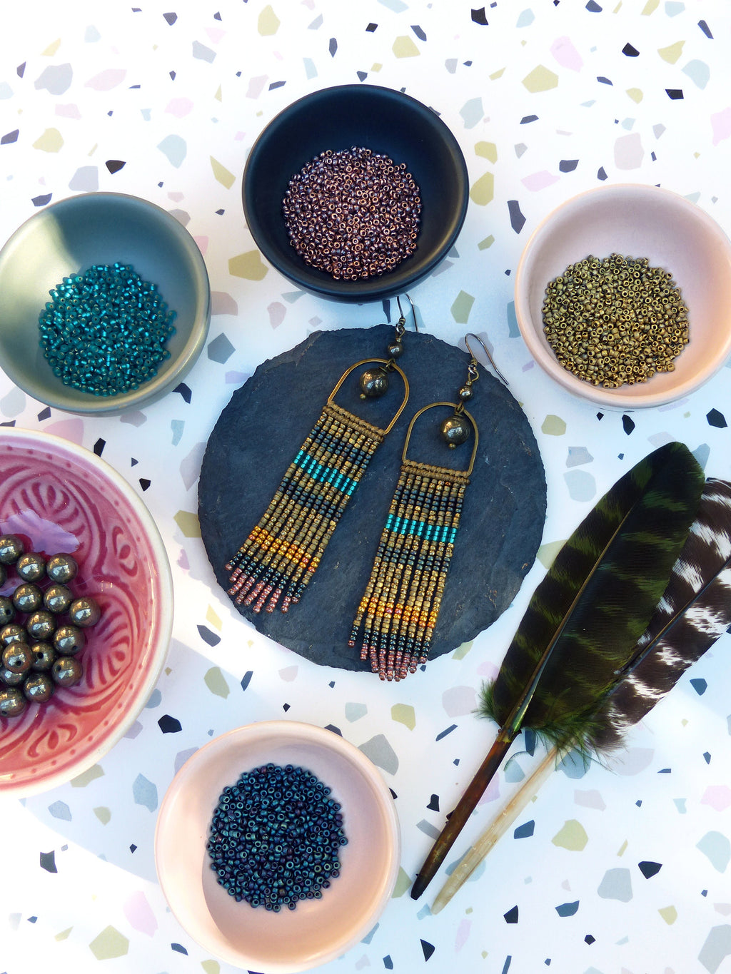 Peut inclure: Une paire de longues boucles d'oreilles en perles avec un anneau en métal doré et une frange de perles colorées. Les boucles d'oreilles sont présentées sur une dalle de schiste noire avec de petits bols de perles de différentes couleurs.