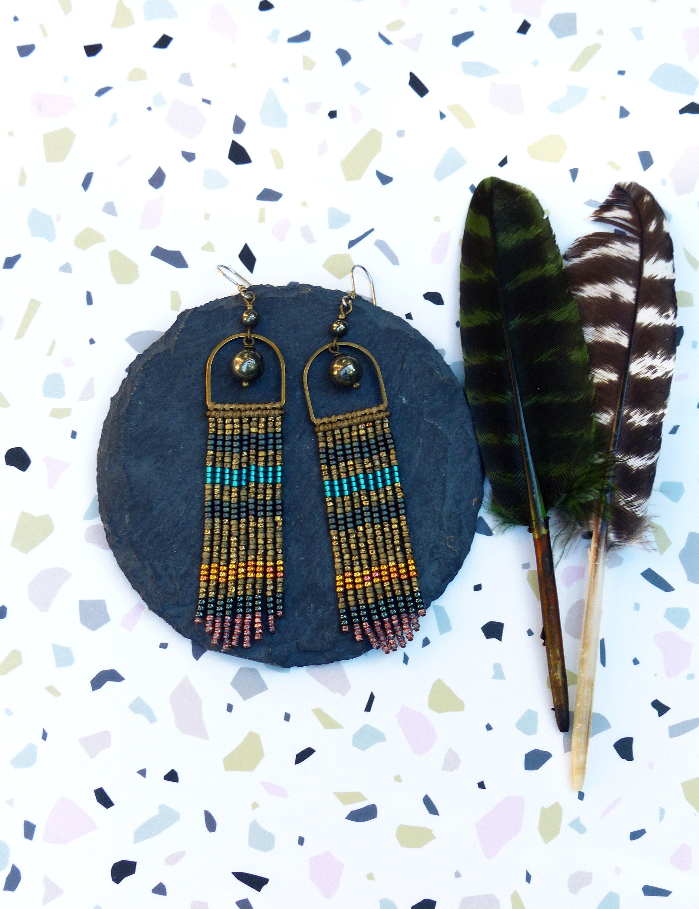 Peut inclure: Une paire de longues boucles d'oreilles en perles avec un motif de couleur or et noir. Les boucles d'oreilles présentent une petite pierre ronde gris foncé au sommet et pendent d'une arche dorée.