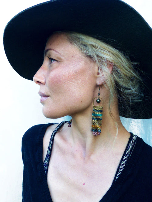 Peut inclure: Une femme portant un chapeau noir et un haut noir avec un détail de fermeture éclair argentée. Elle porte de longues boucles d'oreilles en perles avec un mélange de perles dorées, bleues et vertes.