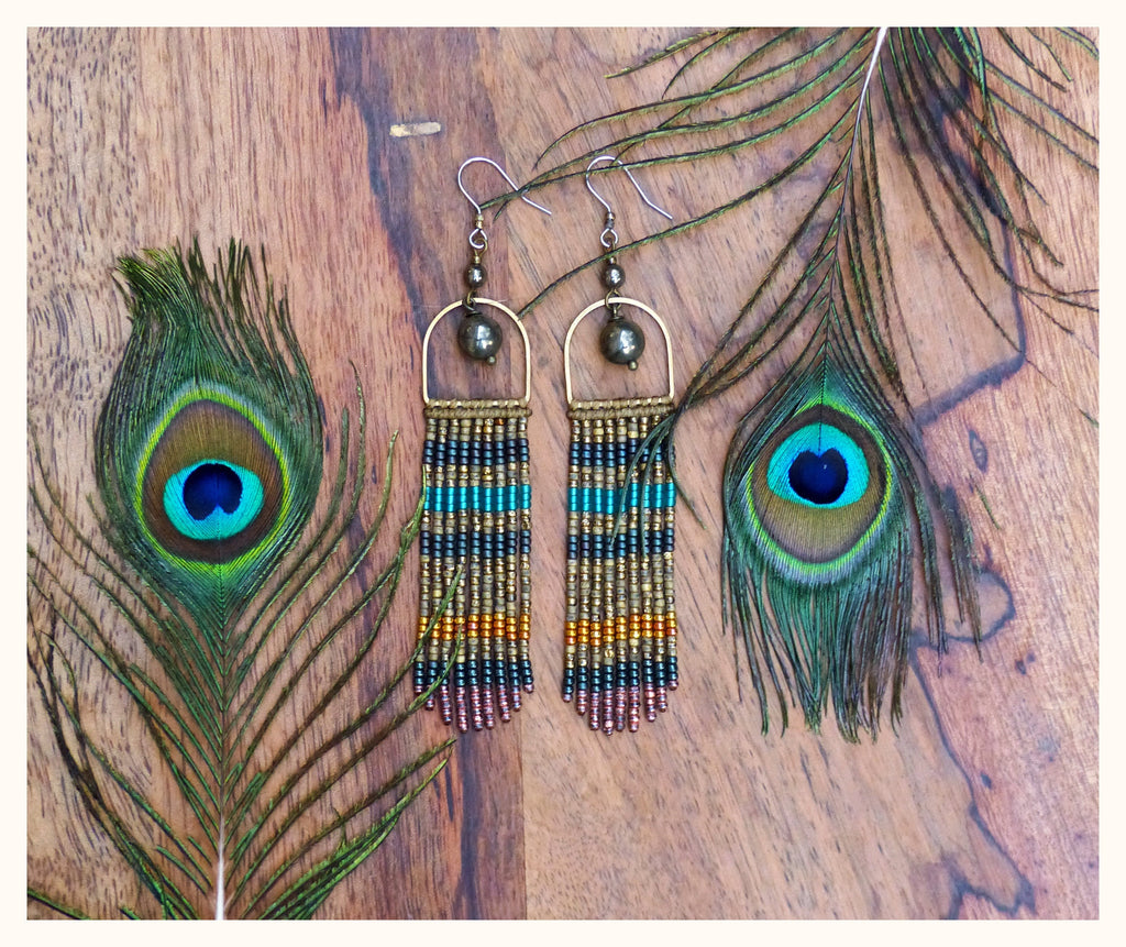 Peut inclure: Une paire de longues boucles d'oreilles en perles avec un cadre doré et un accent de pierre gris foncé. Les boucles d'oreilles sont suspendues sur un fond en bois avec des plumes de paon.