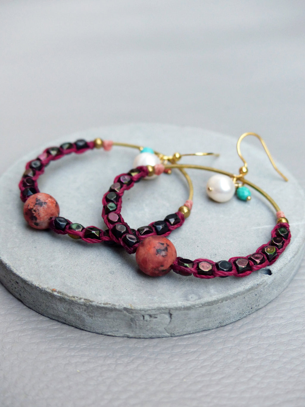 Peut inclure: Une paire de boucles d'oreilles créoles avec un cordon bordeaux, des perles noires et des perles rouges et blanches. Les boucles d'oreilles ont un crochet doré et une perle de pierre rouge.