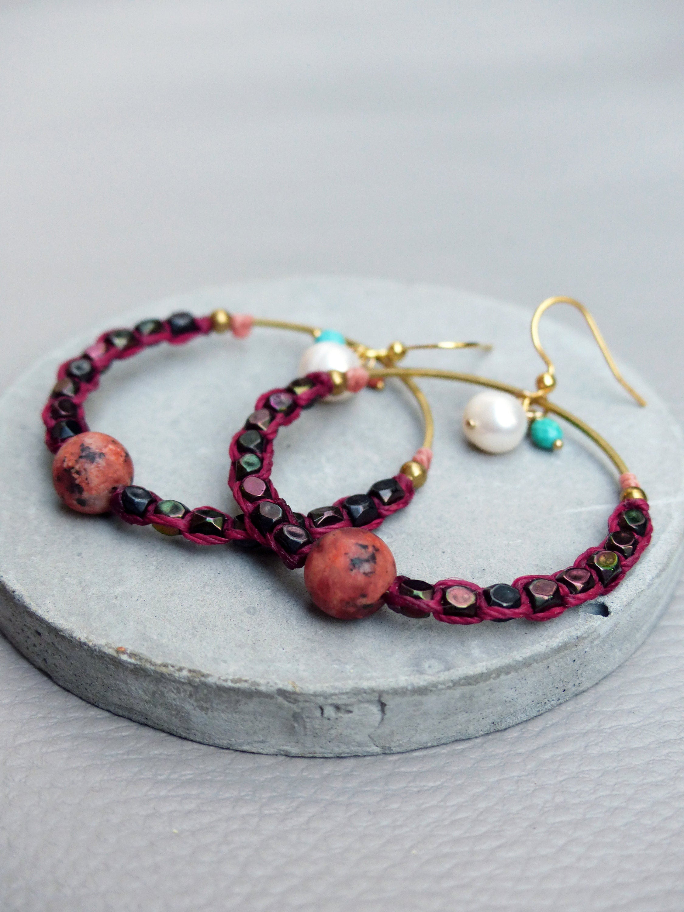 Peut inclure: Une paire de boucles d'oreilles créoles avec un cordon bordeaux, des perles noires et des perles rouges et blanches. Les boucles d'oreilles ont un crochet doré et une perle de pierre rouge.