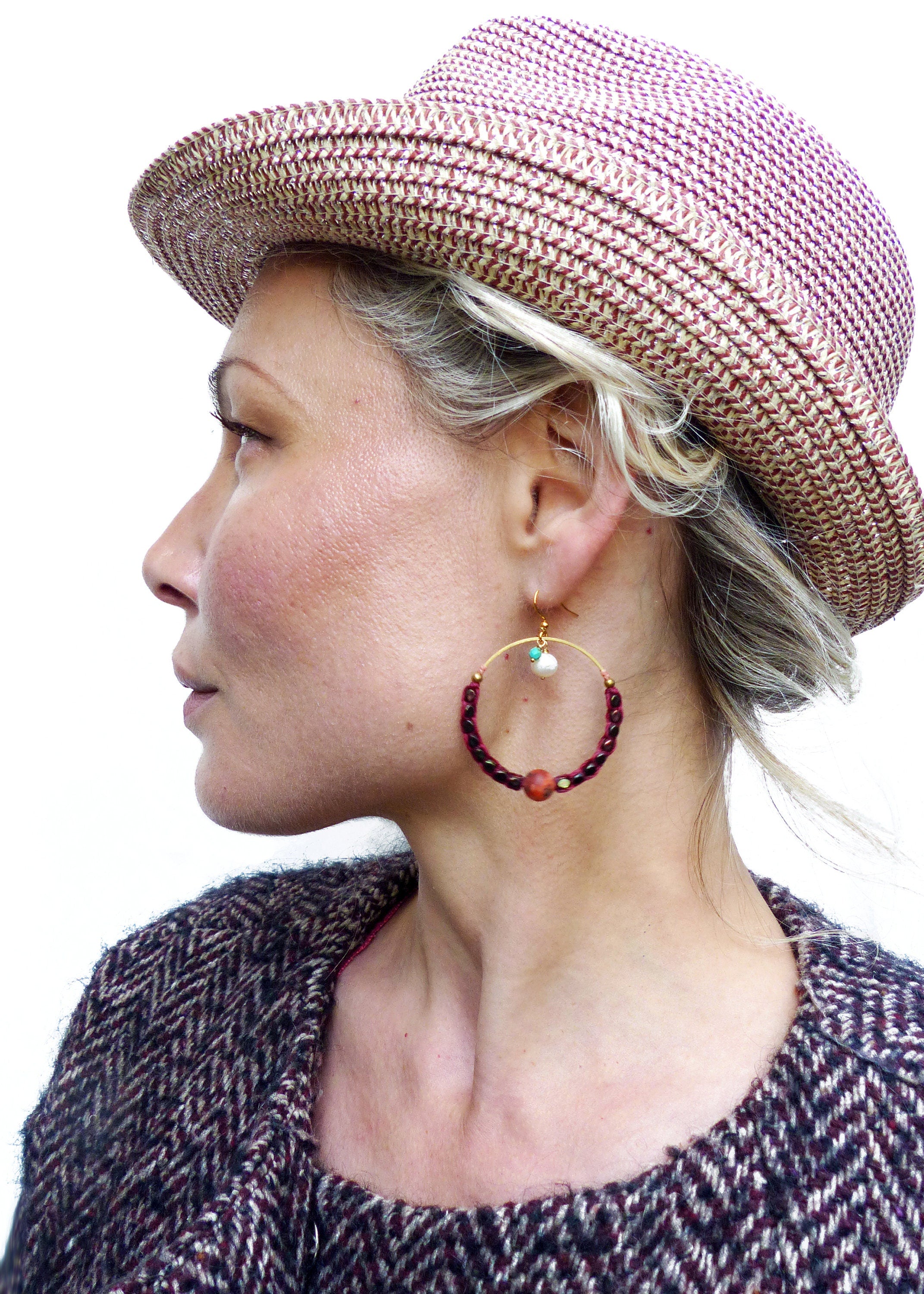 Peut inclure: Une femme portant un chapeau de paille rose et blanc et une veste en tweed marron et blanc. Elle porte de grandes boucles d'oreilles créoles avec des perles rouges, une perle blanche et une perle turquoise.