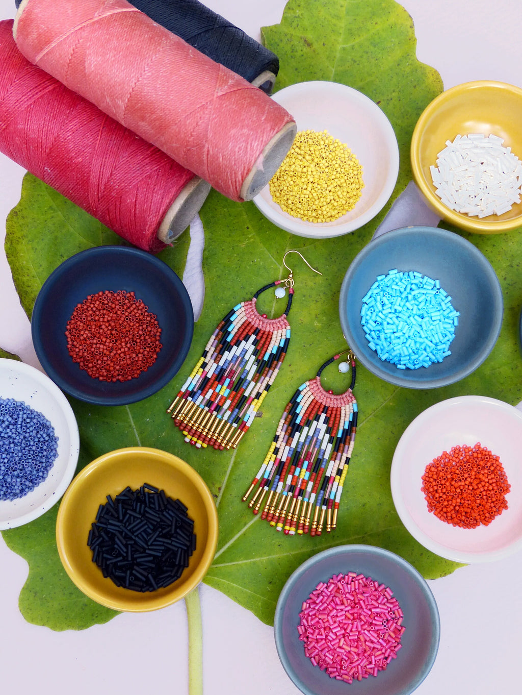 Peut inclure: Une paire de boucles d'oreilles en perles colorées avec des accents dorés, disposées sur une feuille verte avec de petits bols de perles de différentes couleurs, notamment rouge, bleu, blanc, noir et jaune.