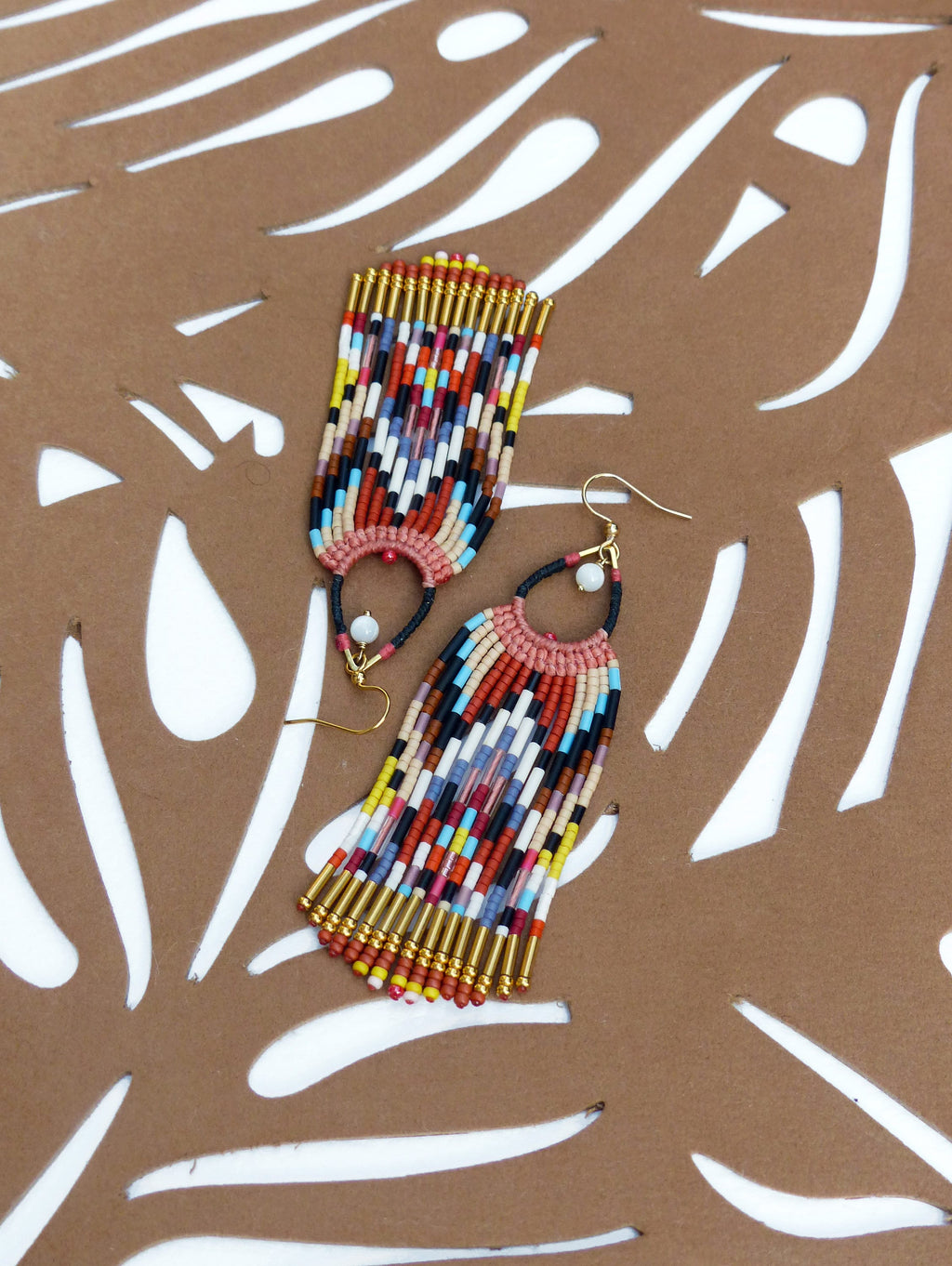 Peut inclure: Une paire de boucles d'oreilles en perles colorées avec des accents dorés. Les boucles d'oreilles sont longues et pendantes, avec un motif géométrique de perles dans des tons de rouge, orange, jaune, bleu, vert et blanc.