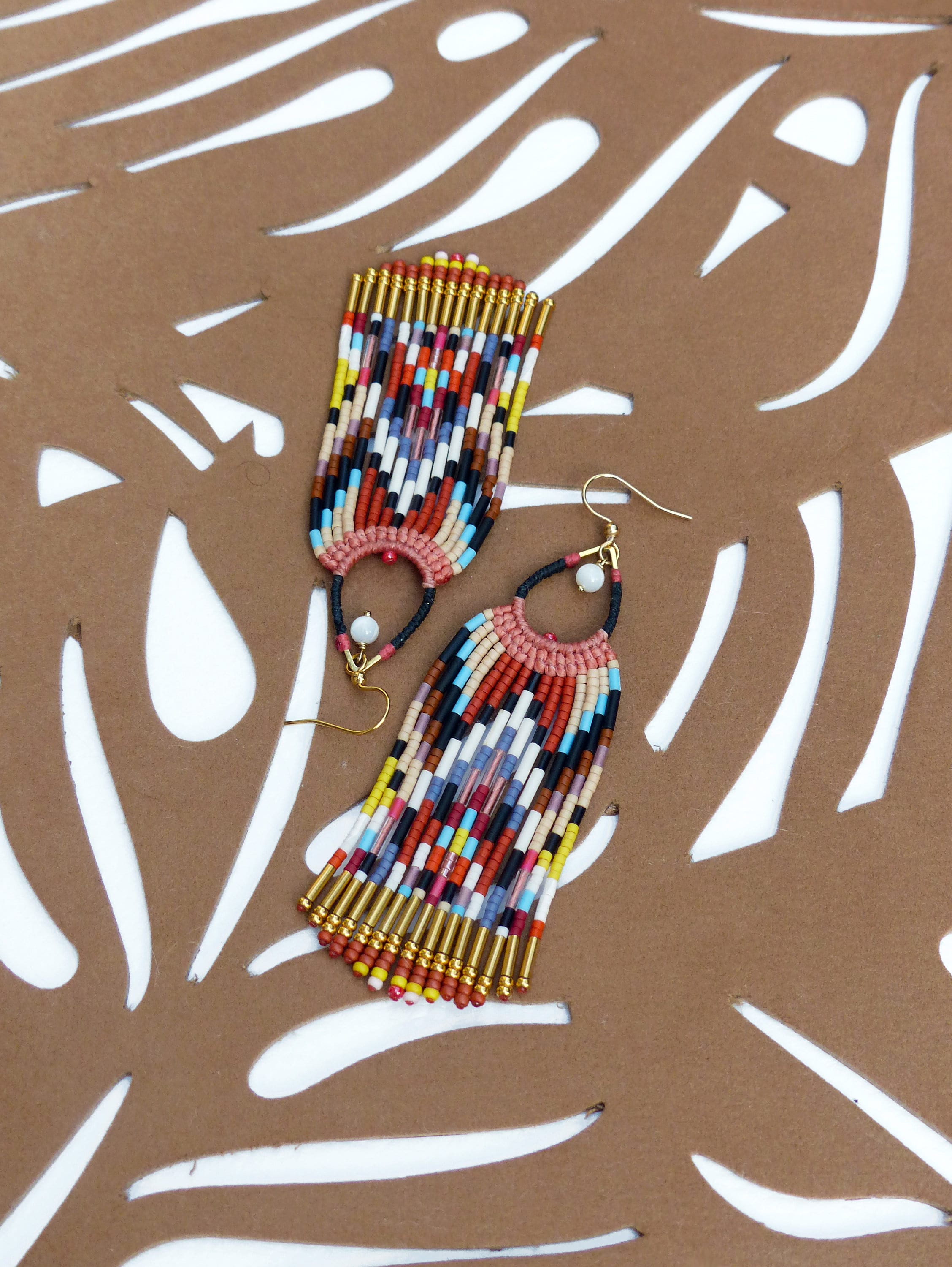 Peut inclure: Une paire de boucles d'oreilles en perles colorées avec des accents dorés. Les boucles d'oreilles sont longues et pendantes, avec un motif géométrique de perles dans des tons de rouge, orange, jaune, bleu, vert et blanc.
