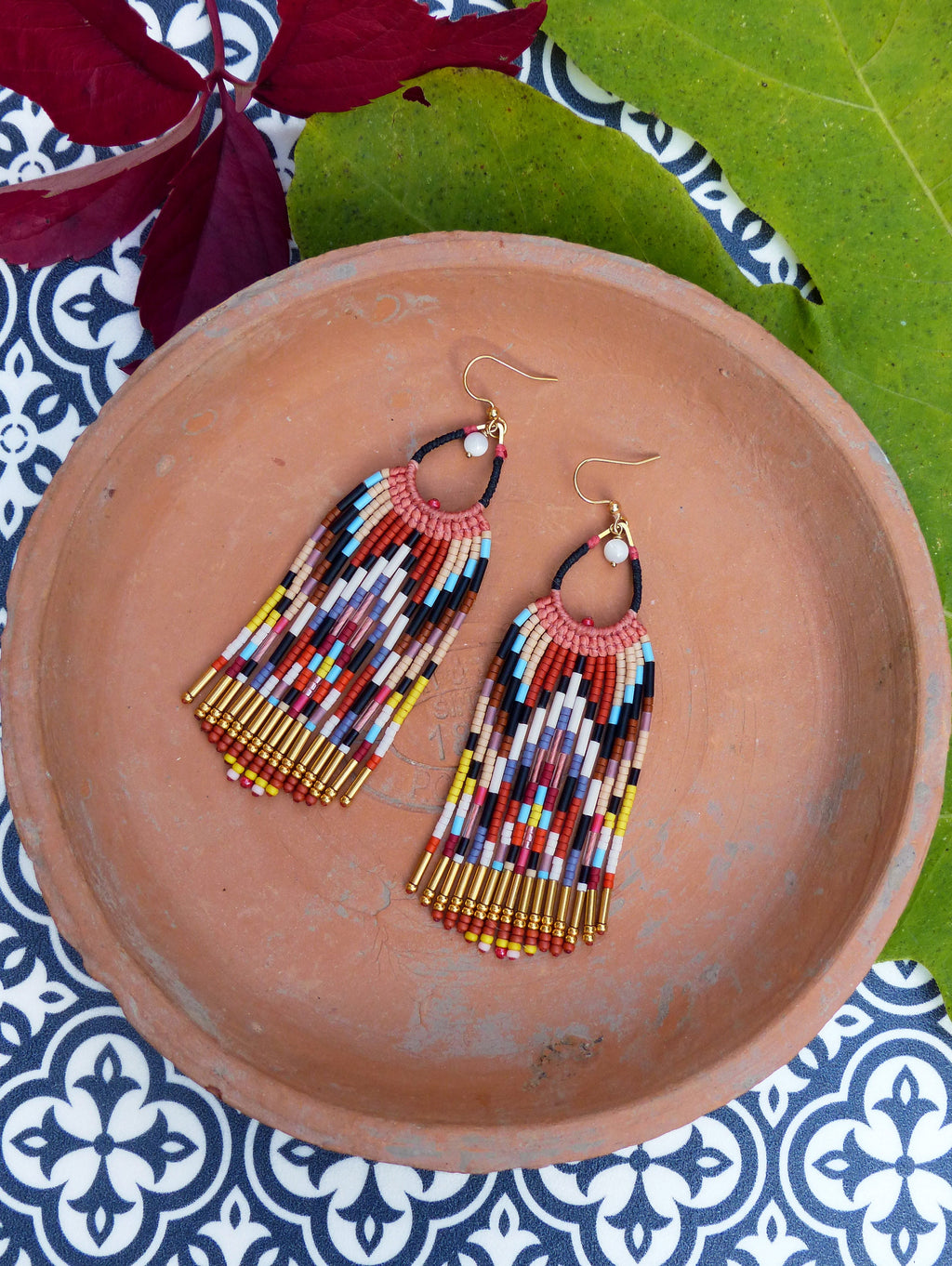 Peut inclure: Une paire de longues boucles d'oreilles en perles colorées avec des accents dorés. Les boucles d'oreilles sont suspendues à une assiette en terre cuite.