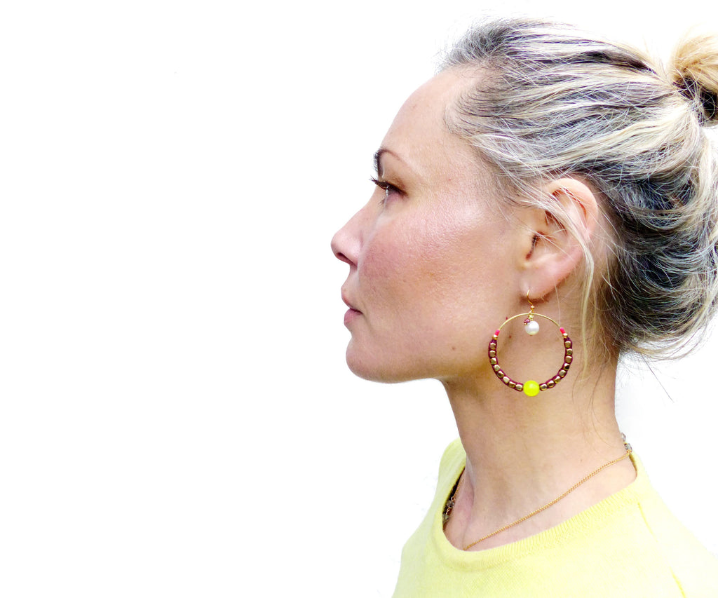Peut inclure: Une paire de boucles d'oreilles créoles avec des perles brunes, une petite perle blanche et une perle jaune vif. Les boucles d'oreilles sont portées par une personne aux cheveux blonds.