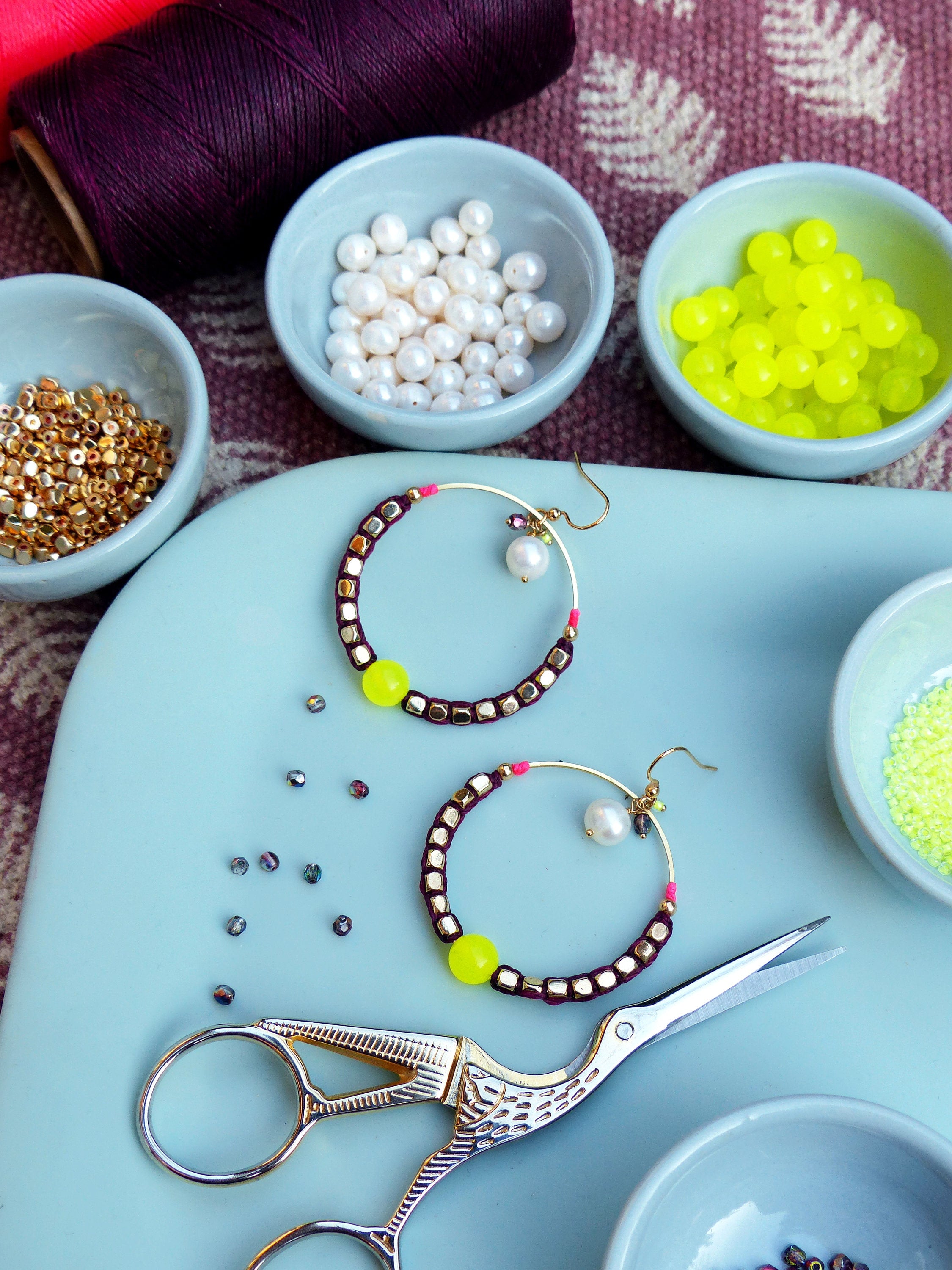 Peut inclure: Une paire de boucles d'oreilles créoles avec un design de perles bordeaux et dorées, une perle d'accent rose et jaune et un charme de perle. Les boucles d'oreilles sont sur une surface bleu clair avec une paire de ciseaux argentés et de petits bols de perles.