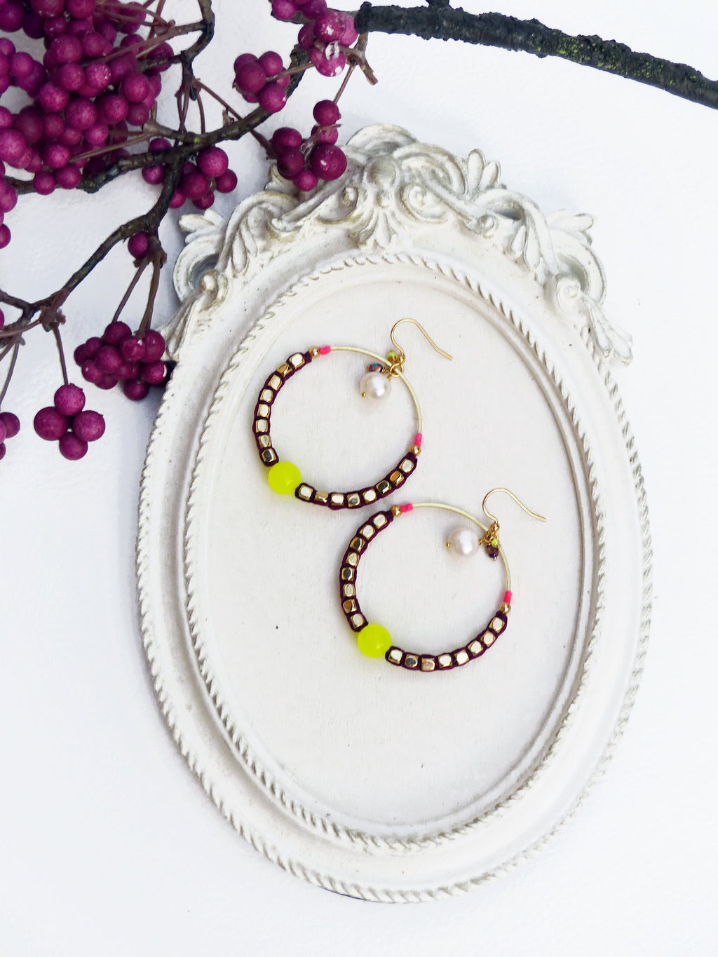 Peut inclure: Une paire de boucles d'oreilles créoles avec un design de perles bordeaux et dorées. Chaque boucle d'oreille est ornée d'un petit charme de perle blanche et d'une perle jaune vif.