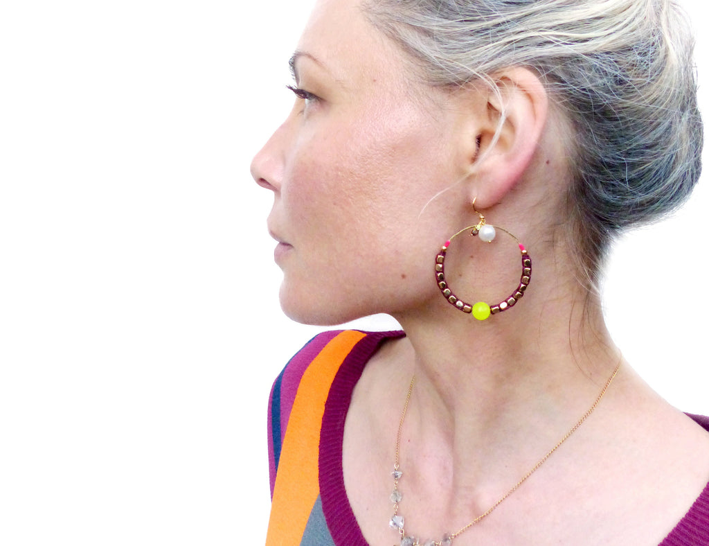 Peut inclure: Une paire de boucles d'oreilles créoles avec une base en fil d'or et des perles bordeaux. Une petite perle jaune et une perle blanche pendent du cercle.
