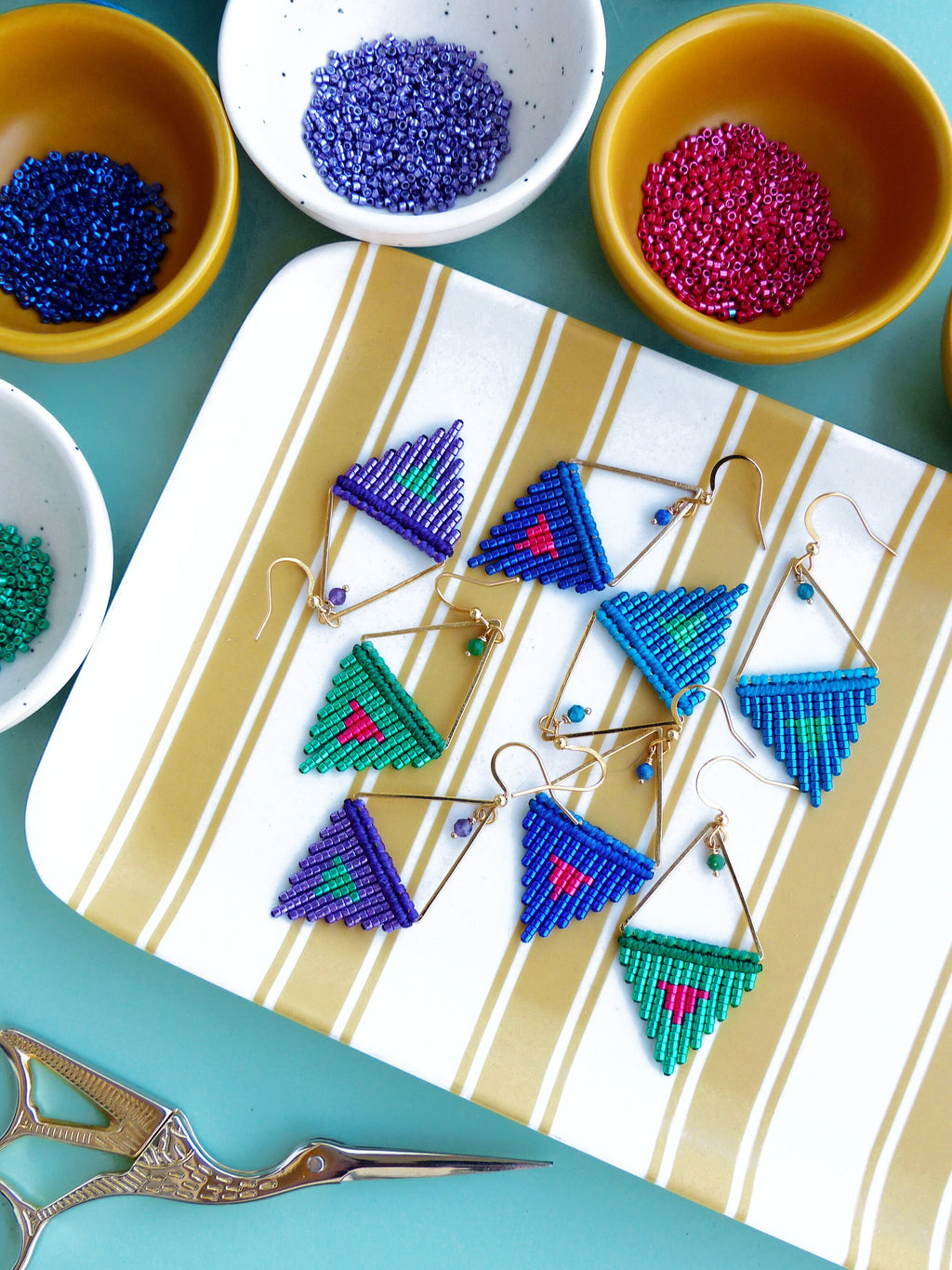 Peut inclure: Boucles d'oreilles macramé faites à la main, de différentes couleurs, notamment bleu, vert et violet, avec des montures en métal doré. Les boucles d'oreilles sont présentées sur un plateau à rayures, avec des petits bols de perles en arrière-plan. Une paire de ciseaux est également visible.