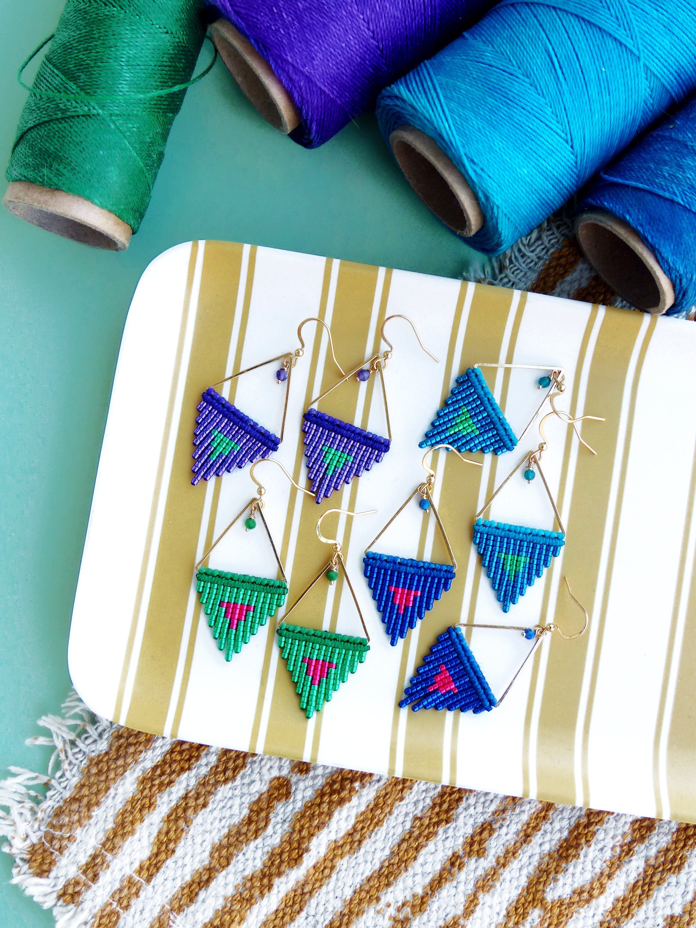 Peut inclure: Boucles d'oreilles macramé faites à la main, de différentes couleurs, notamment vert, bleu et violet. Les boucles d'oreilles ont une forme triangulaire avec des montures en métal doré et des accents de perles. Elles sont présentées sur un plateau rayé.
