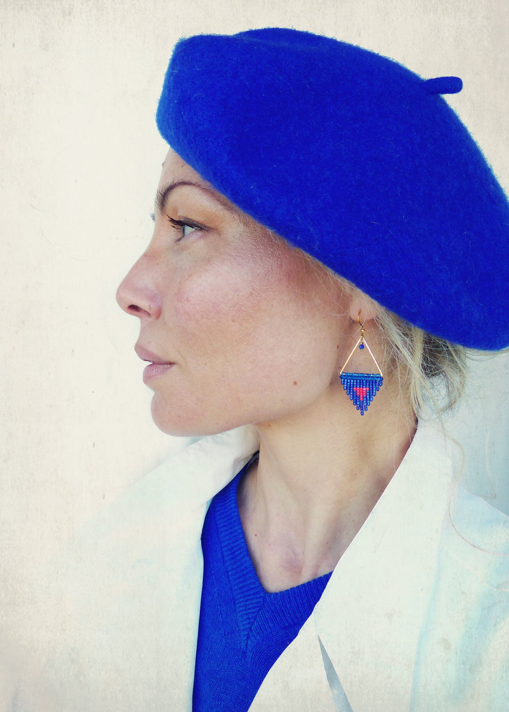 Peut inclure: Une femme porte un béret bleu roi et un manteau blanc. Elle porte également des boucles d'oreilles triangulaires dorées avec un motif de perles bleues et rouges. Les boucles d'oreilles sont de forme géométrique avec un cœur au centre.