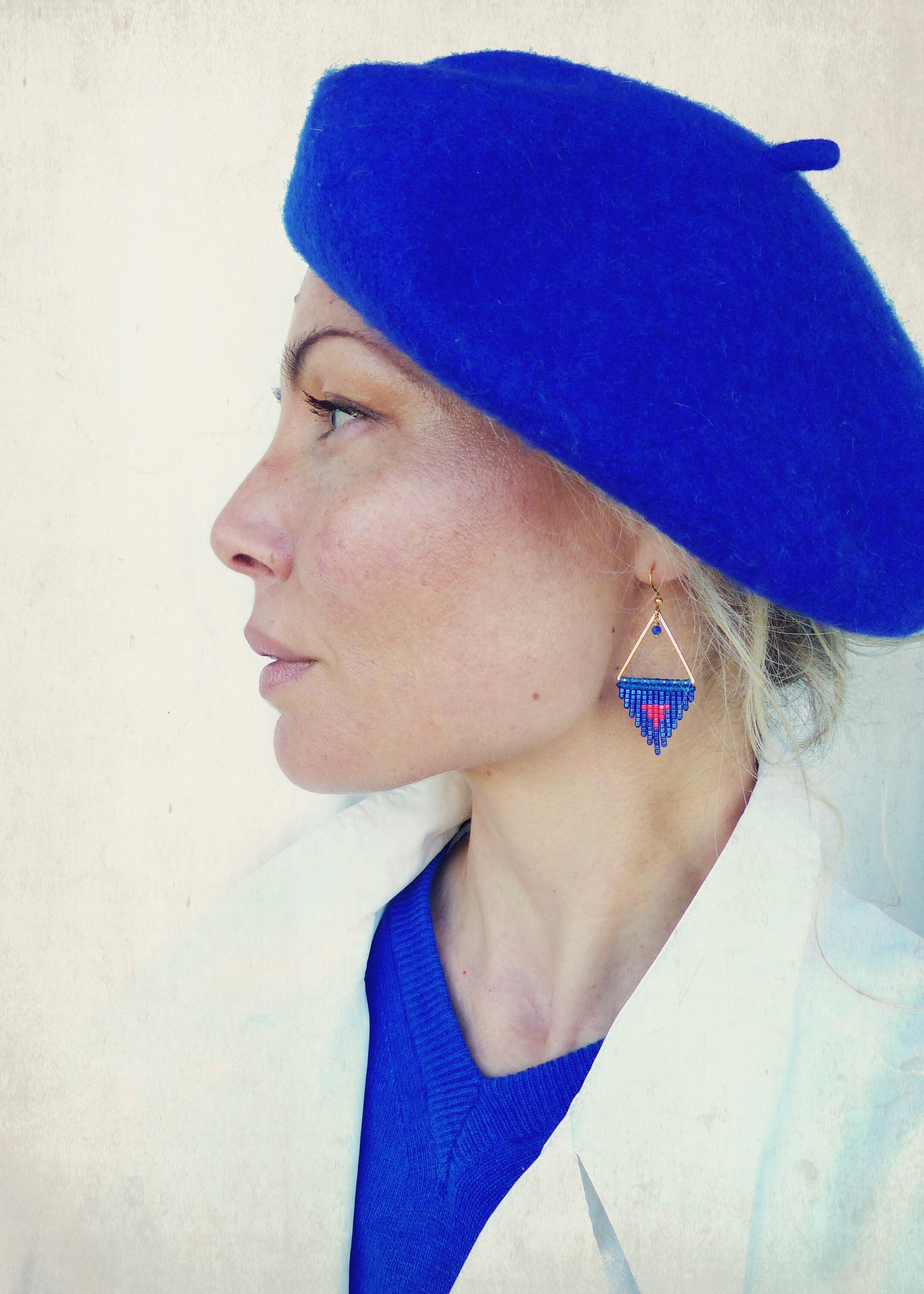 Peut inclure: Une femme porte un béret bleu roi et un manteau blanc. Elle porte également des boucles d'oreilles triangulaires dorées avec un motif de perles bleues et rouges. Les boucles d'oreilles sont de forme géométrique avec un cœur au centre.