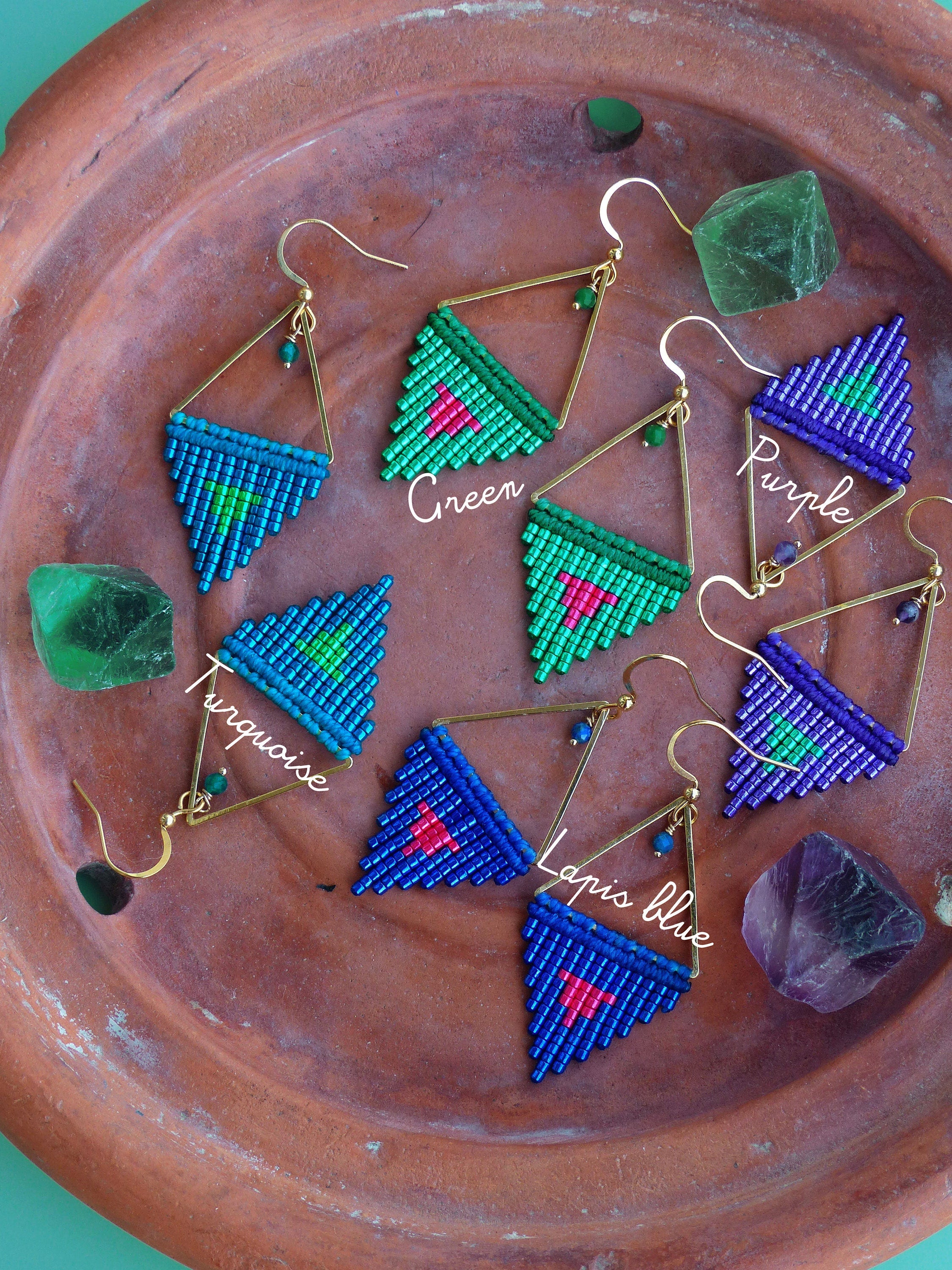 Peut inclure: Boucles d'oreilles macramé faites à la main, disponibles en turquoise, vert, violet et bleu lapis. Chaque boucle d'oreille présente un motif triangulaire avec des accents métalliques dorés et des crochets d'oreille. Présentées sur une surface couleur terre cuite.