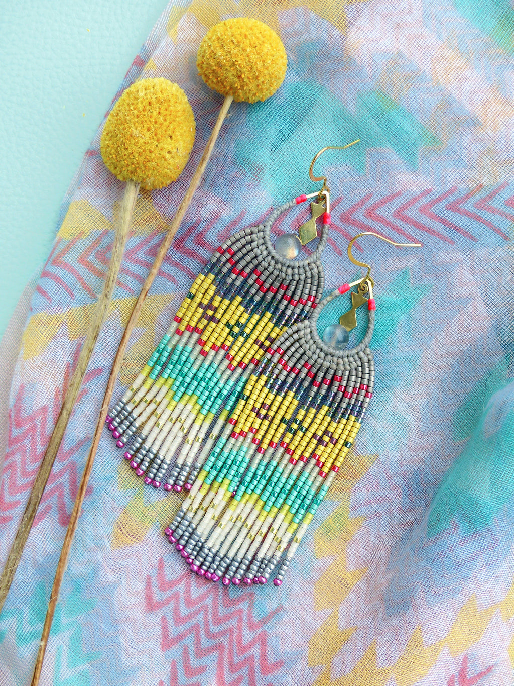 Peut inclure: Une paire de longues boucles d'oreilles en perles avec un design en frange. Les boucles d'oreilles sont faites de perles grises, blanches, jaunes, bleues et roses. Les boucles d'oreilles sont suspendues à des crochets dorés.