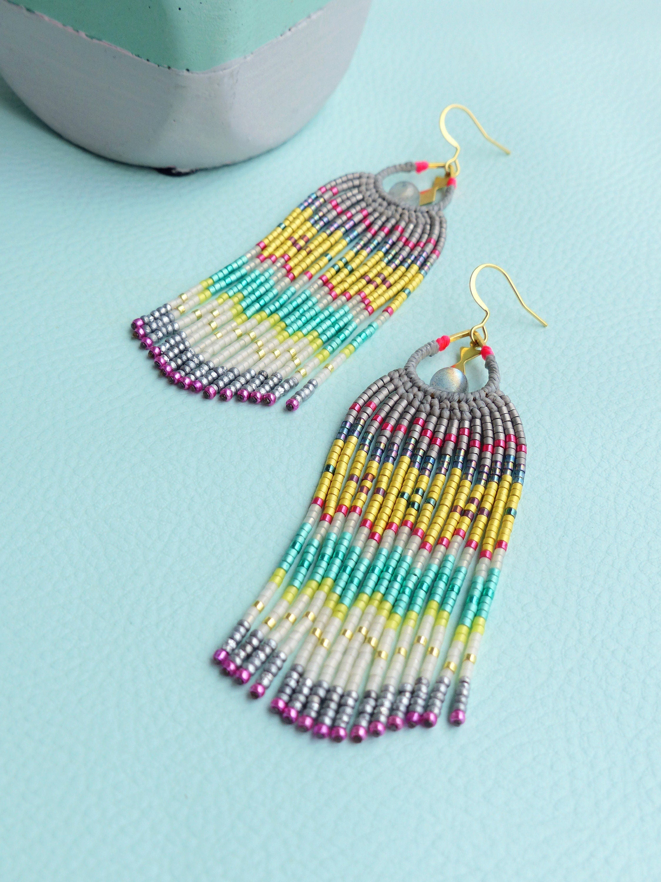 Peut inclure: Une paire de longues boucles d'oreilles en perles colorées avec un anneau gris et or en haut. Les boucles d'oreilles sont faites de petites perles colorées dans des tons de rose, jaune, vert et argent.