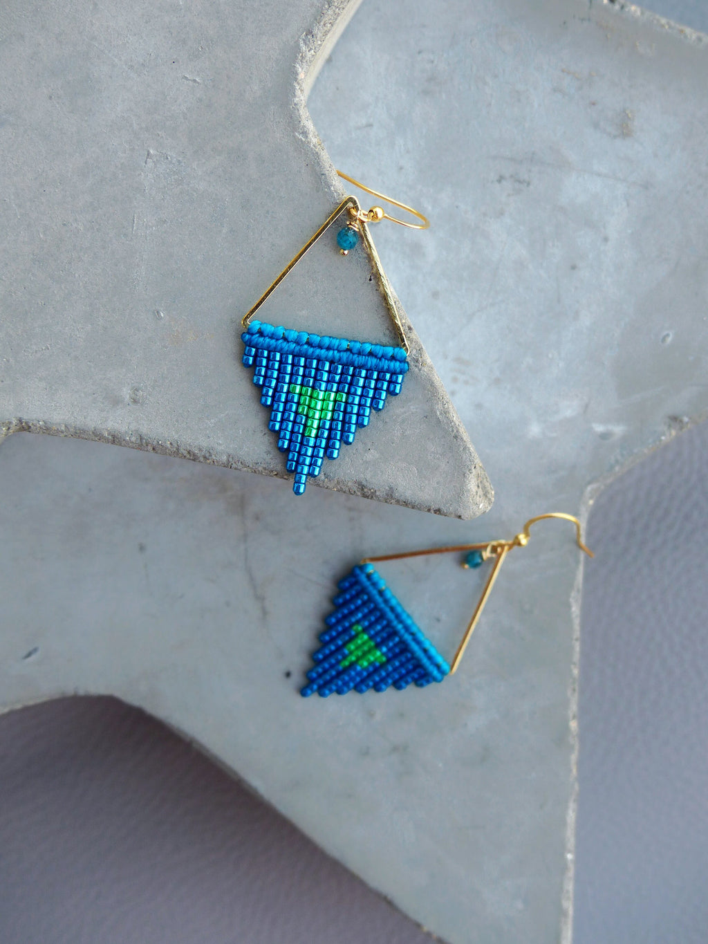 Peut inclure: Paire de boucles d'oreilles faites à la main avec un motif géométrique. Les boucles d'oreilles ont un cadre doré en forme de triangle. Le triangle est rempli de perles bleues et vertes. Fermeture à crochet.