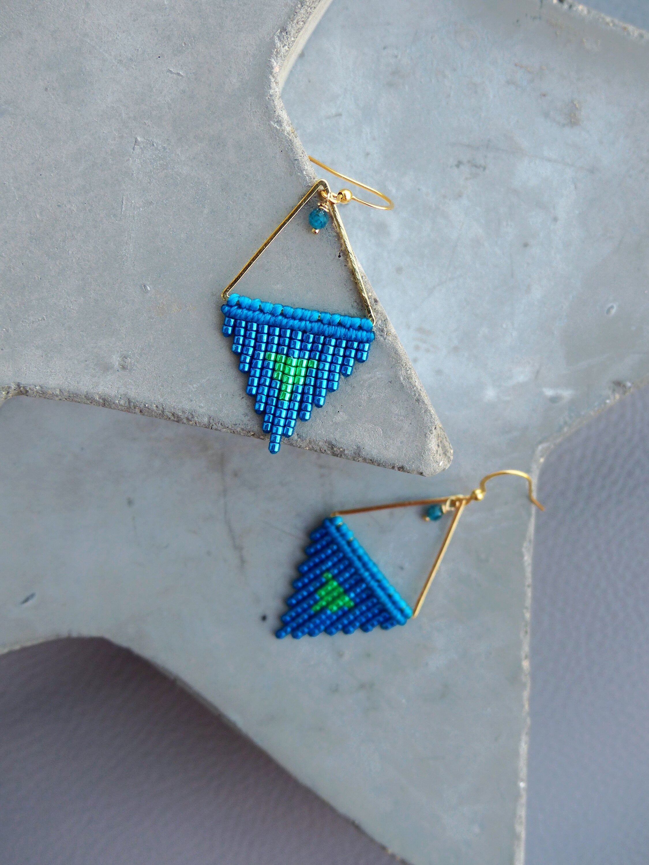 Peut inclure: Paire de boucles d'oreilles faites à la main avec un motif géométrique. Les boucles d'oreilles ont un cadre doré en forme de triangle. Le triangle est rempli de perles bleues et vertes. Fermeture à crochet.