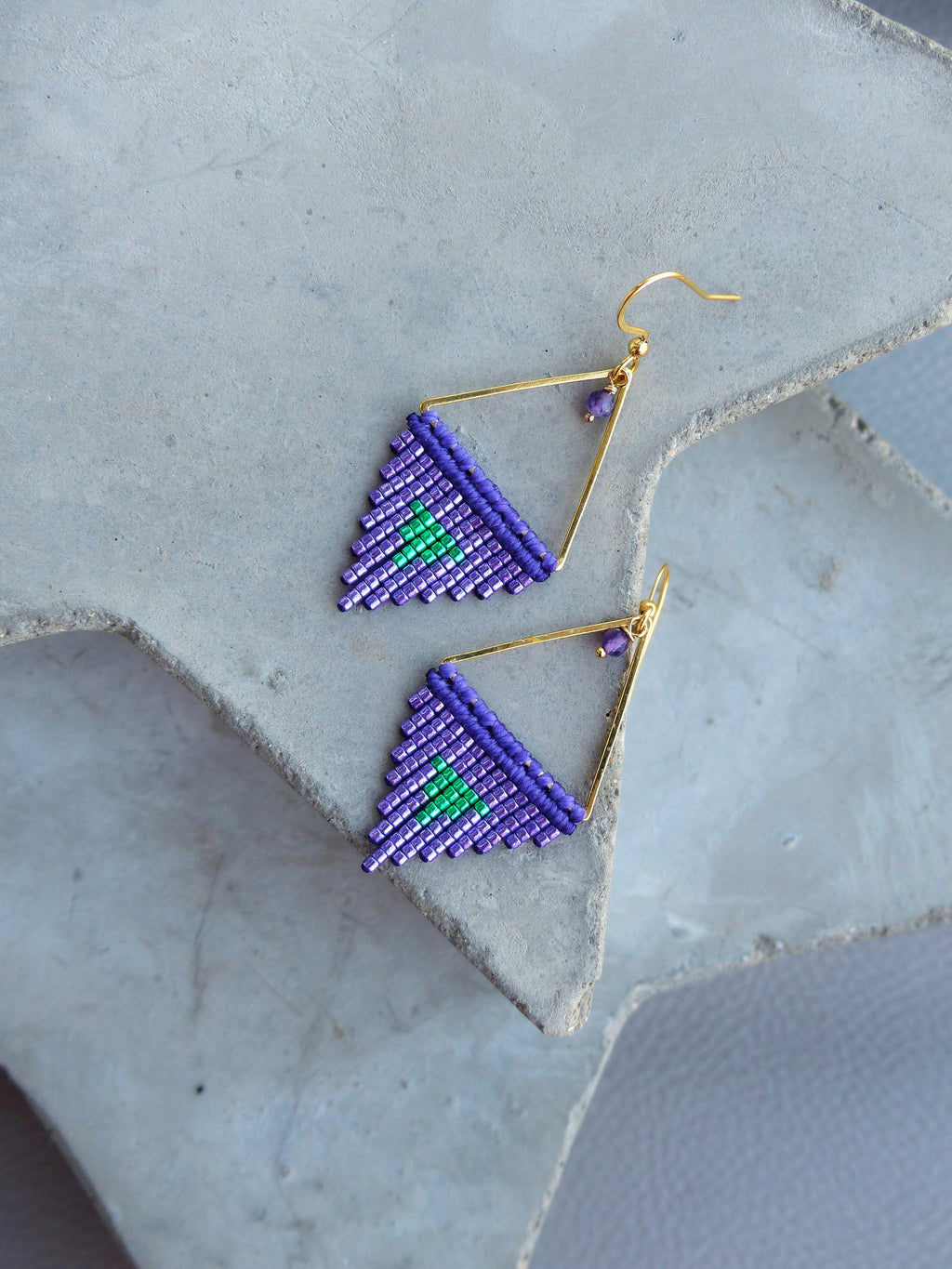 Peut inclure: Une paire de boucles d'oreilles faites à la main avec un motif géométrique. Les boucles d'oreilles présentent un cadre doré avec un triangle perlé dans des tons de violet et de vert. Les boucles d'oreilles sont présentées sur une surface grise.