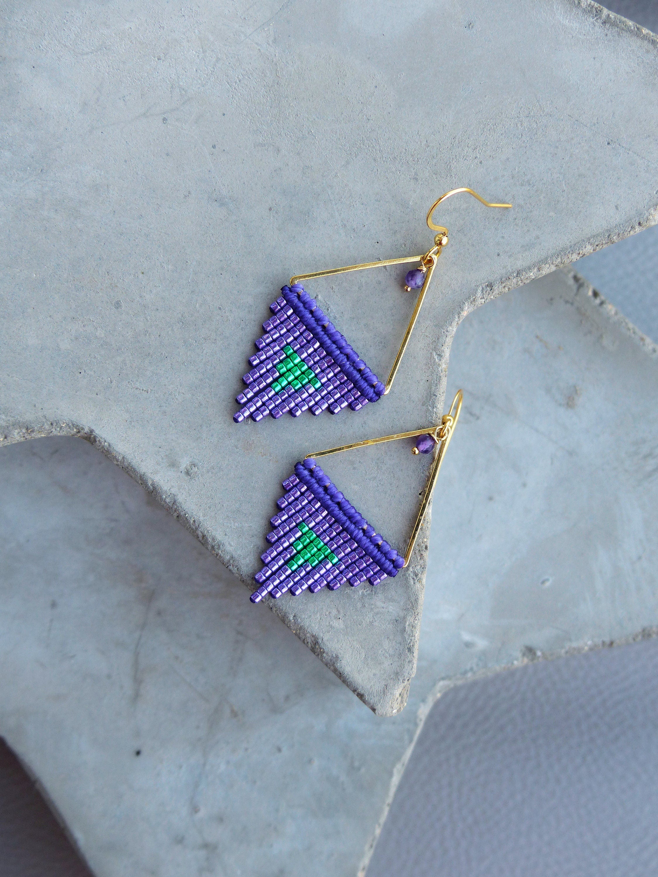 Peut inclure: Une paire de boucles d'oreilles faites à la main avec un motif géométrique. Les boucles d'oreilles présentent un cadre doré avec un triangle perlé dans des tons de violet et de vert. Les boucles d'oreilles sont présentées sur une surface grise.