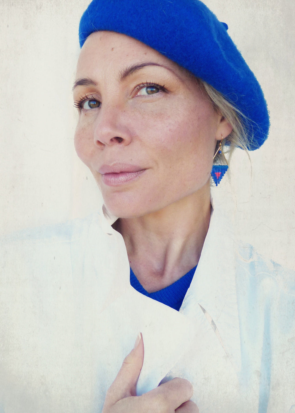 Peut inclure: Une femme porte un béret bleu et des boucles d'oreilles dorées avec un motif triangulaire. Les boucles d'oreilles comportent des perles rouges et bleues. Elle porte une veste blanche et un pull bleu.
