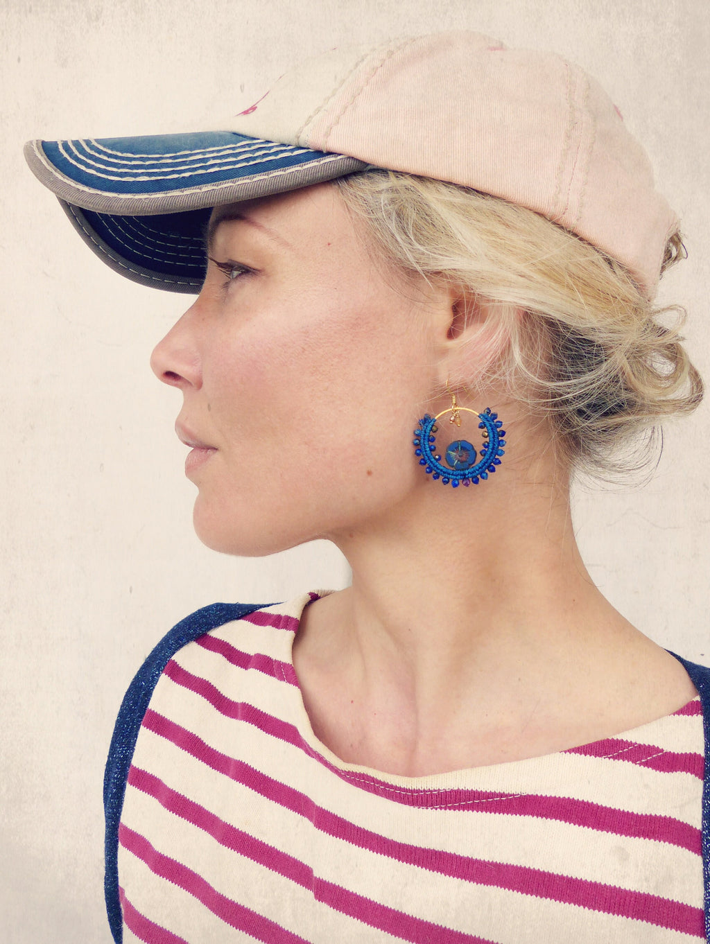 Peut inclure: Une femme portant une casquette de baseball rose avec une visière bleue et une chemise rayée. Elle porte des boucles d'oreilles créoles en perles bleues.