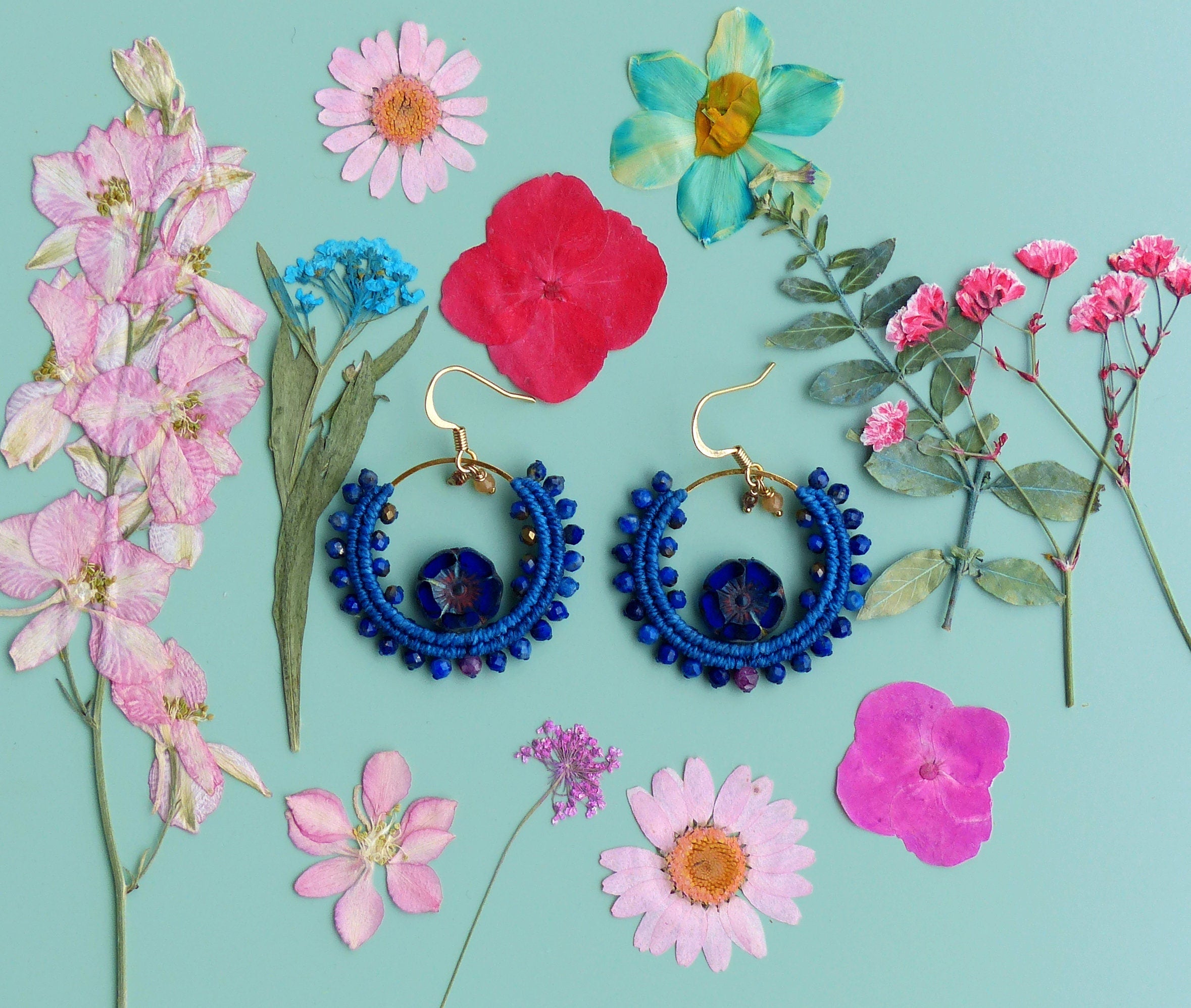 Peut inclure: Une paire de boucles d'oreilles créoles bleues et dorées avec une pierre précieuse bleue au centre. Les boucles d'oreilles sont entourées de fleurs séchées roses, bleues et blanches.