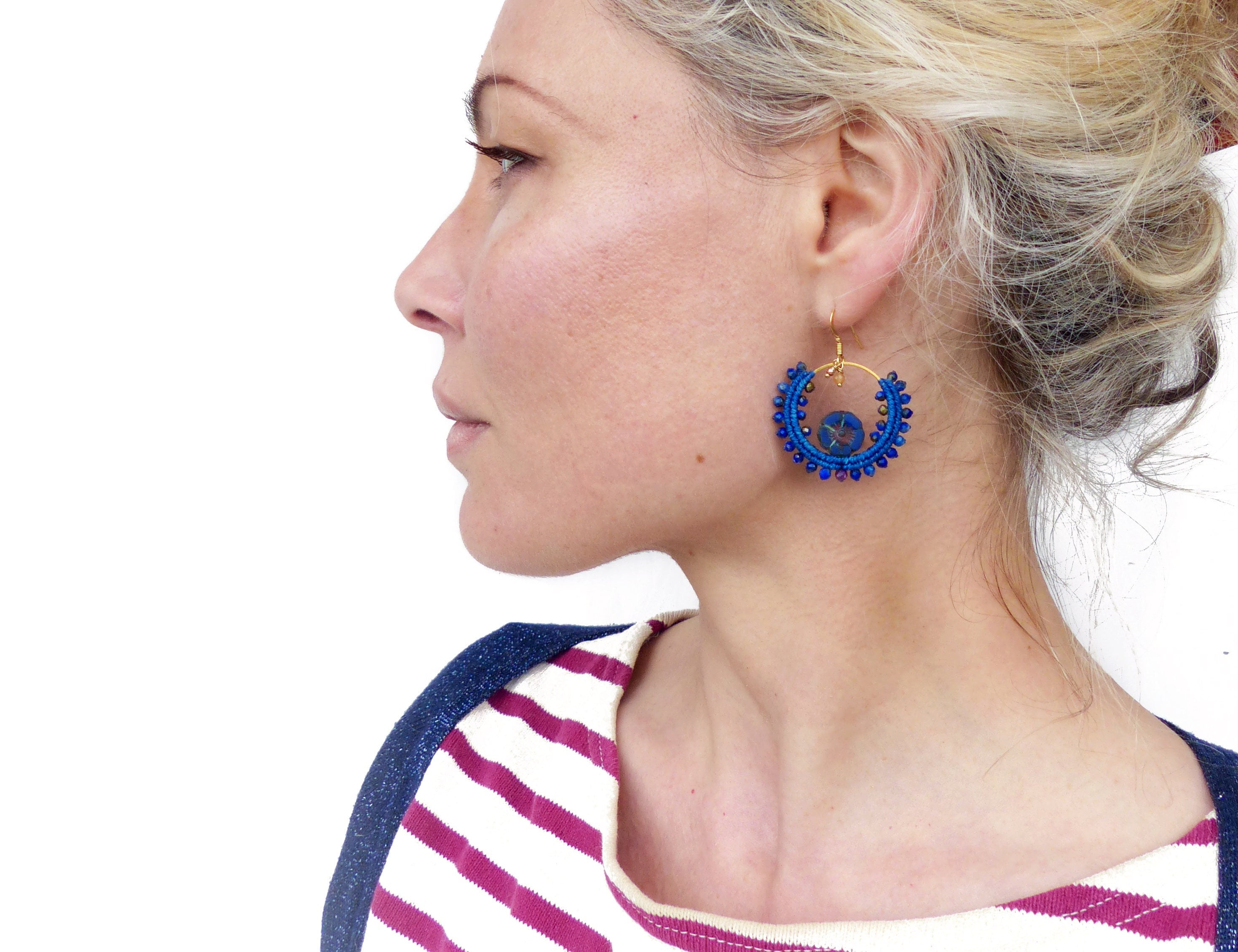 Peut inclure: Une paire de boucles d'oreilles créoles bleues avec un cadre en fil d'or et un motif de perles bleues. Les boucles d'oreilles sont portées par une personne aux cheveux blonds.