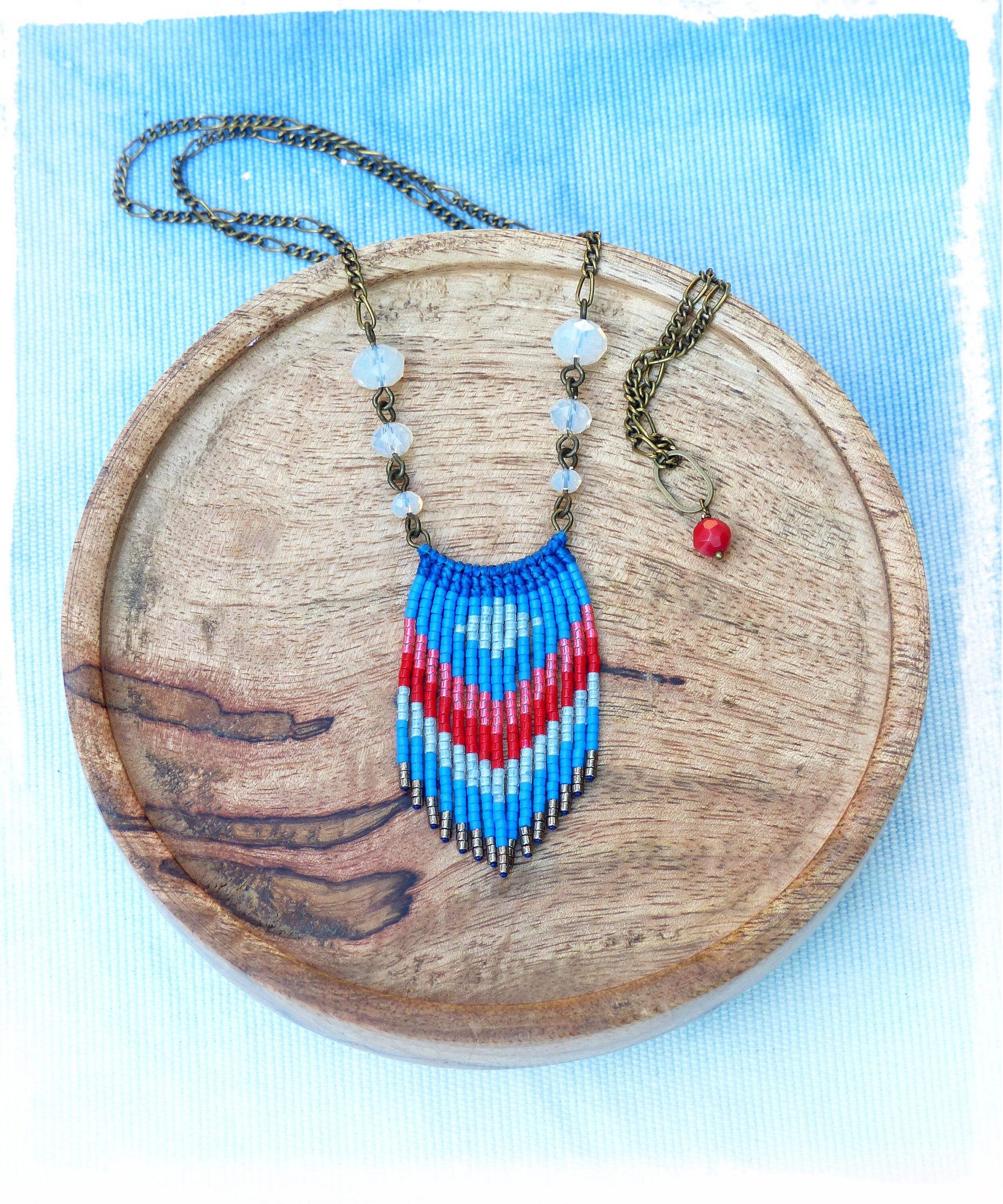 Peut inclure: Un collier macramé fait à la main avec un pendentif à chevrons bleu, rouge et blanc. Le collier est orné d'une chaîne en bronze et de petites perles transparentes. Une petite perle rouge est suspendue à une chaîne séparée. Le collier repose sur un plateau en bois.