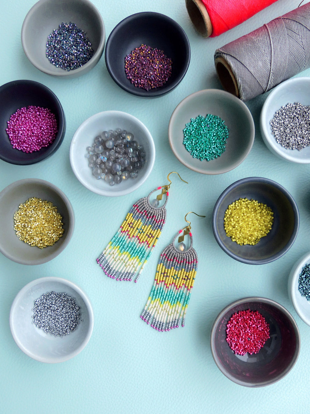 Peut inclure: Une paire de boucles d'oreilles en perles colorées avec un design en frange. Les boucles d'oreilles sont faites de petites perles colorées et ont une monture en métal doré. Les boucles d'oreilles sont présentées sur une surface bleu clair avec de petits bols de perles de différentes couleurs.
