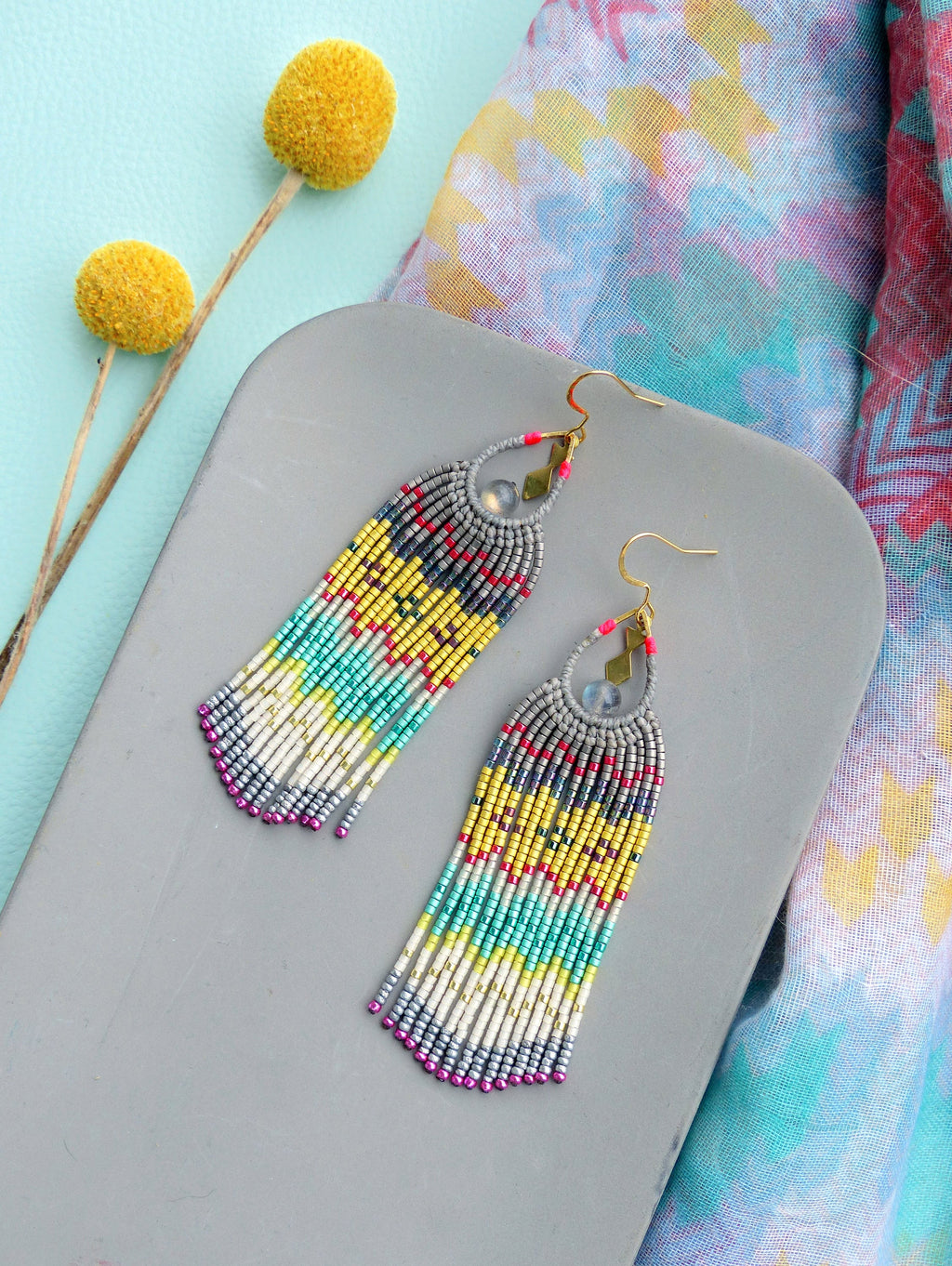 Peut inclure: Une paire de longues boucles d'oreilles en perles colorées avec un schéma de couleurs gris, blanc, jaune, turquoise, rose et rouge. Les boucles d'oreilles ont un crochet doré et une perle argentée au bas.