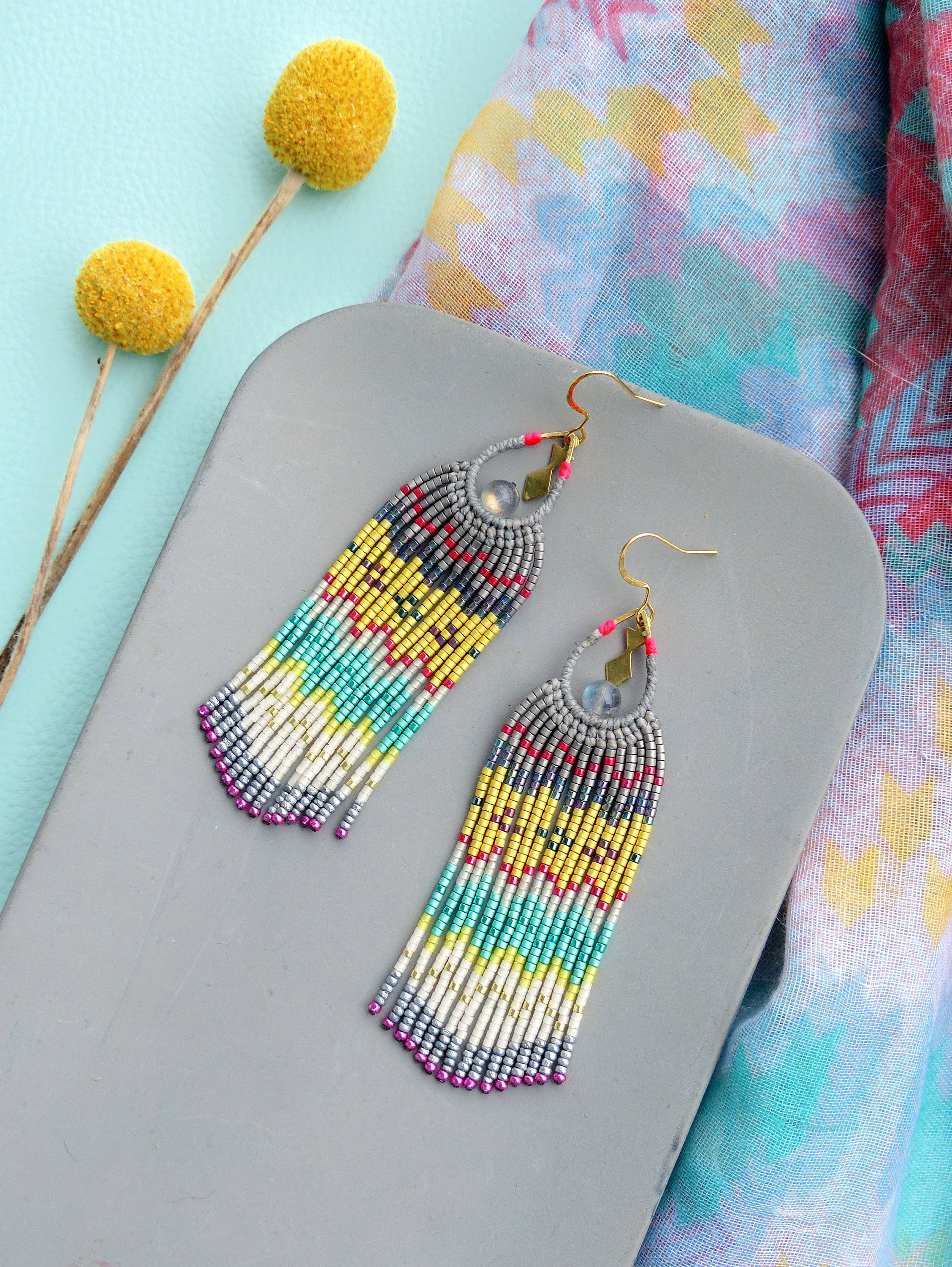 Peut inclure: Une paire de longues boucles d'oreilles en perles colorées avec un schéma de couleurs gris, blanc, jaune, turquoise, rose et rouge. Les boucles d'oreilles ont un crochet doré et une perle argentée au bas.