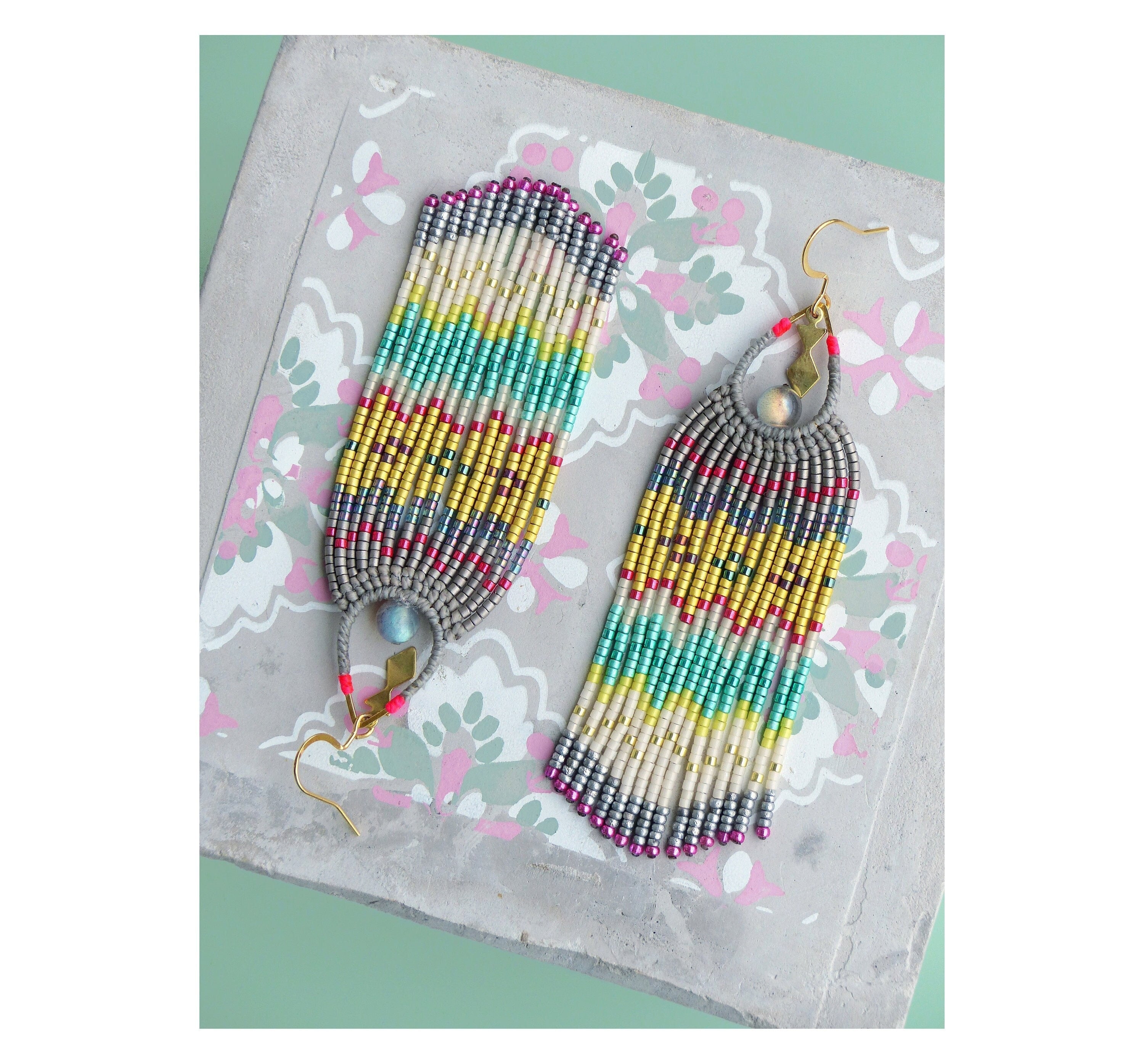 Peut inclure: Une paire de longues boucles d'oreilles en perles colorées avec un motif géométrique. Les boucles d'oreilles sont faites de perles argentées, dorées, blanches, turquoise et roses. Les boucles d'oreilles ont une fermeture à crochet dorée.