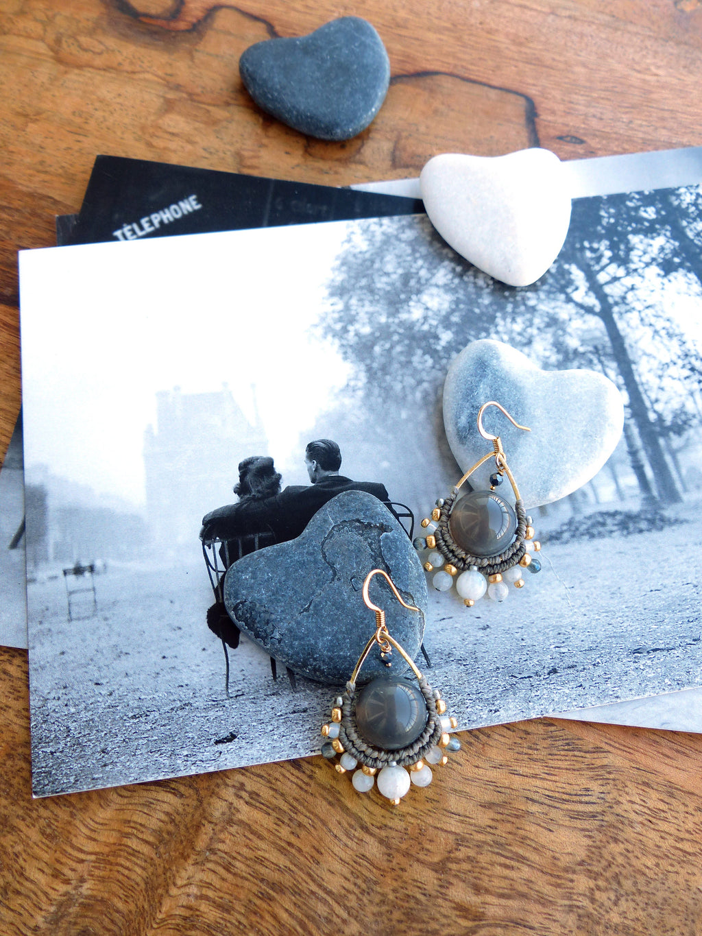 Peut inclure: Boucles d'oreilles grises et dorées faites à la main avec un design en forme de goutte d'eau. Les boucles d'oreilles présentent une grosse perle grise, entourée d'un tissage de style macramé, et agrémentée de petites perles blanches et dorées. Elles sont présentées sur une photographie en noir et blanc.