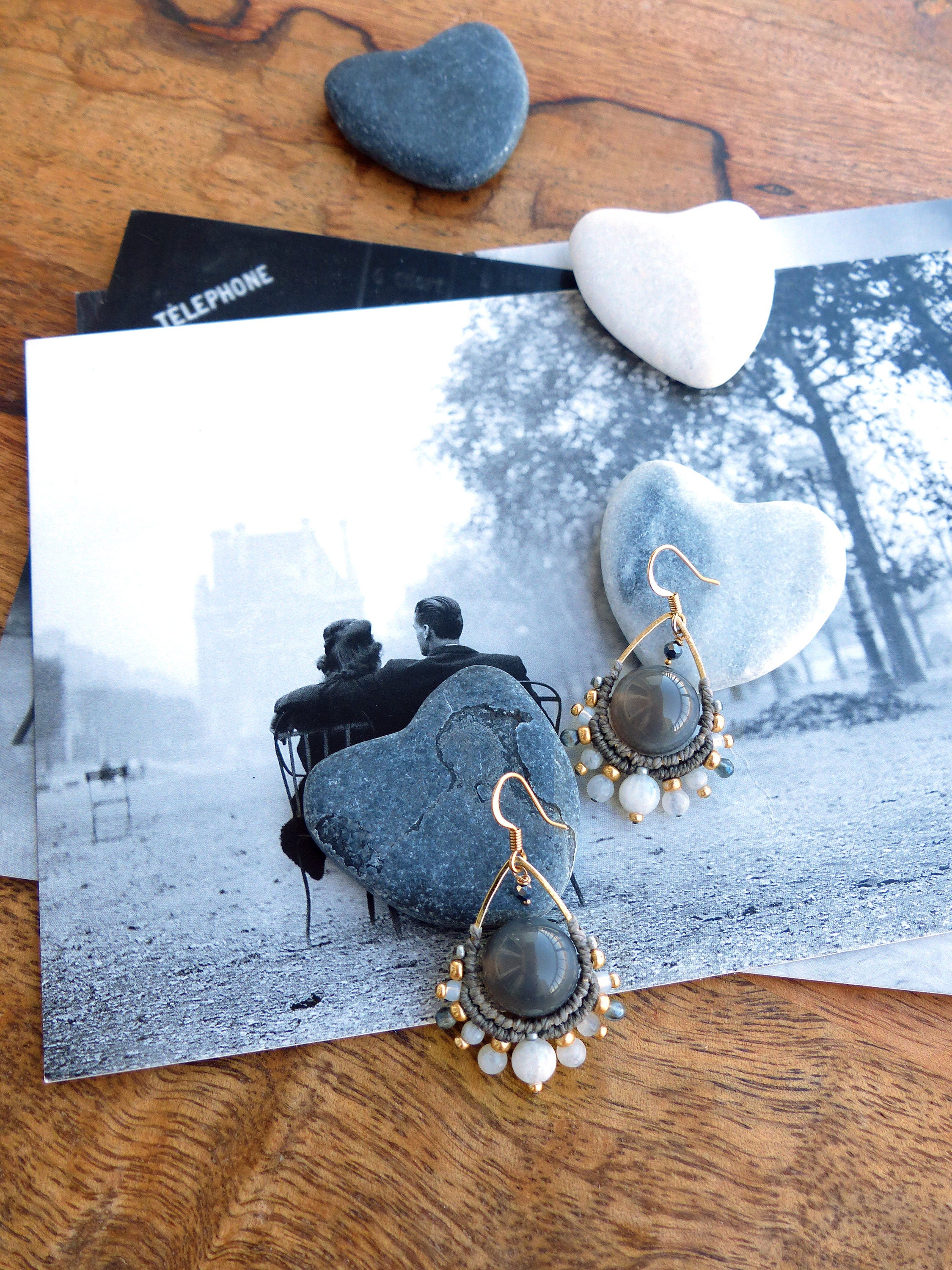Peut inclure: Boucles d'oreilles grises et dorées faites à la main avec un design en forme de goutte d'eau. Les boucles d'oreilles présentent une grosse perle grise, entourée d'un tissage de style macramé, et agrémentée de petites perles blanches et dorées. Elles sont présentées sur une photographie en noir et blanc.