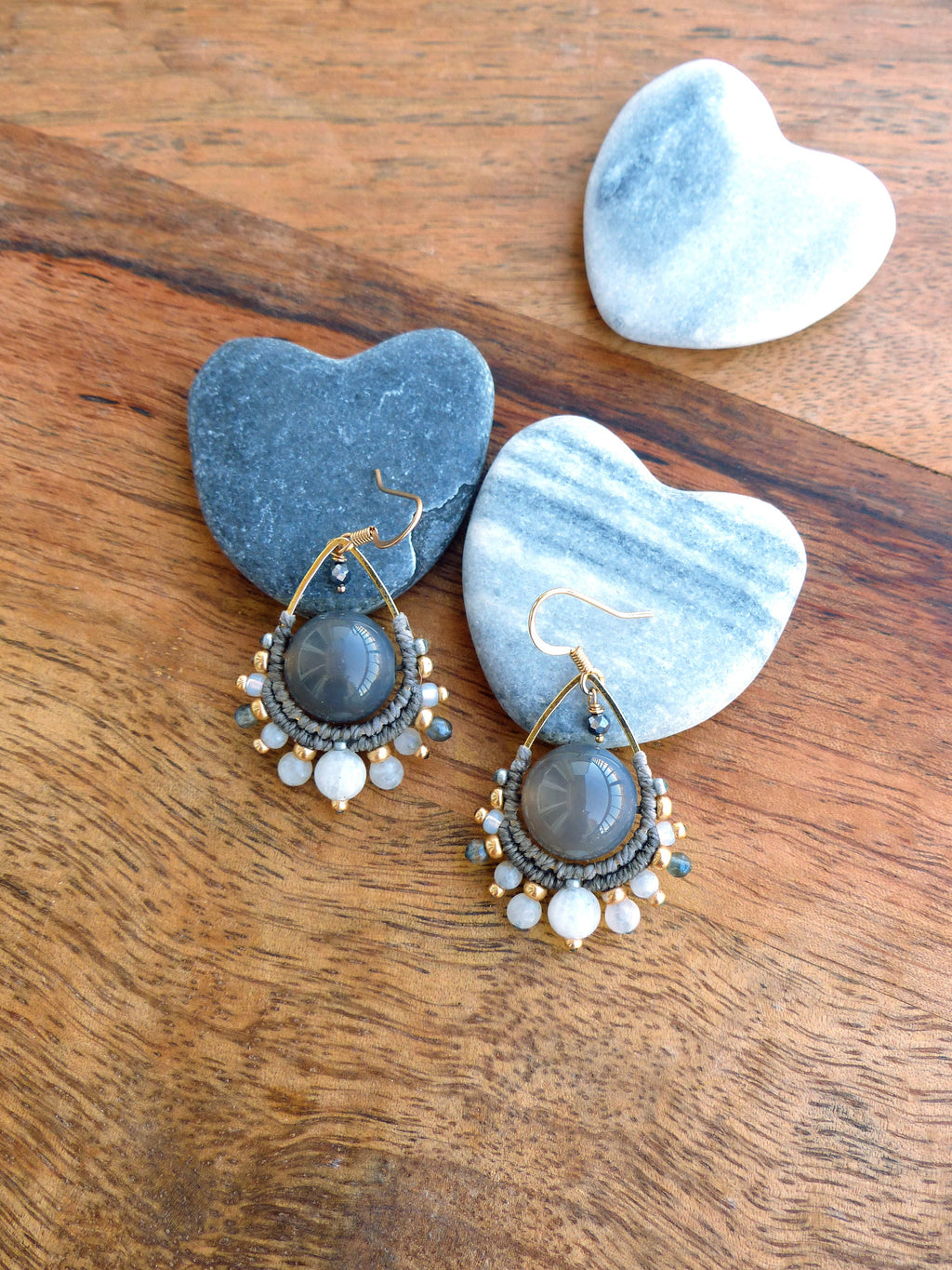 Peut inclure: Boucles d'oreilles artisanales avec une grosse perle grise centrale, entourée d'un motif macramé avec des détails dorés et de petites perles blanches et grises. Les boucles d'oreilles ont des crochets d'oreilles dorés. Des pierres en forme de cœur sont en arrière-plan.