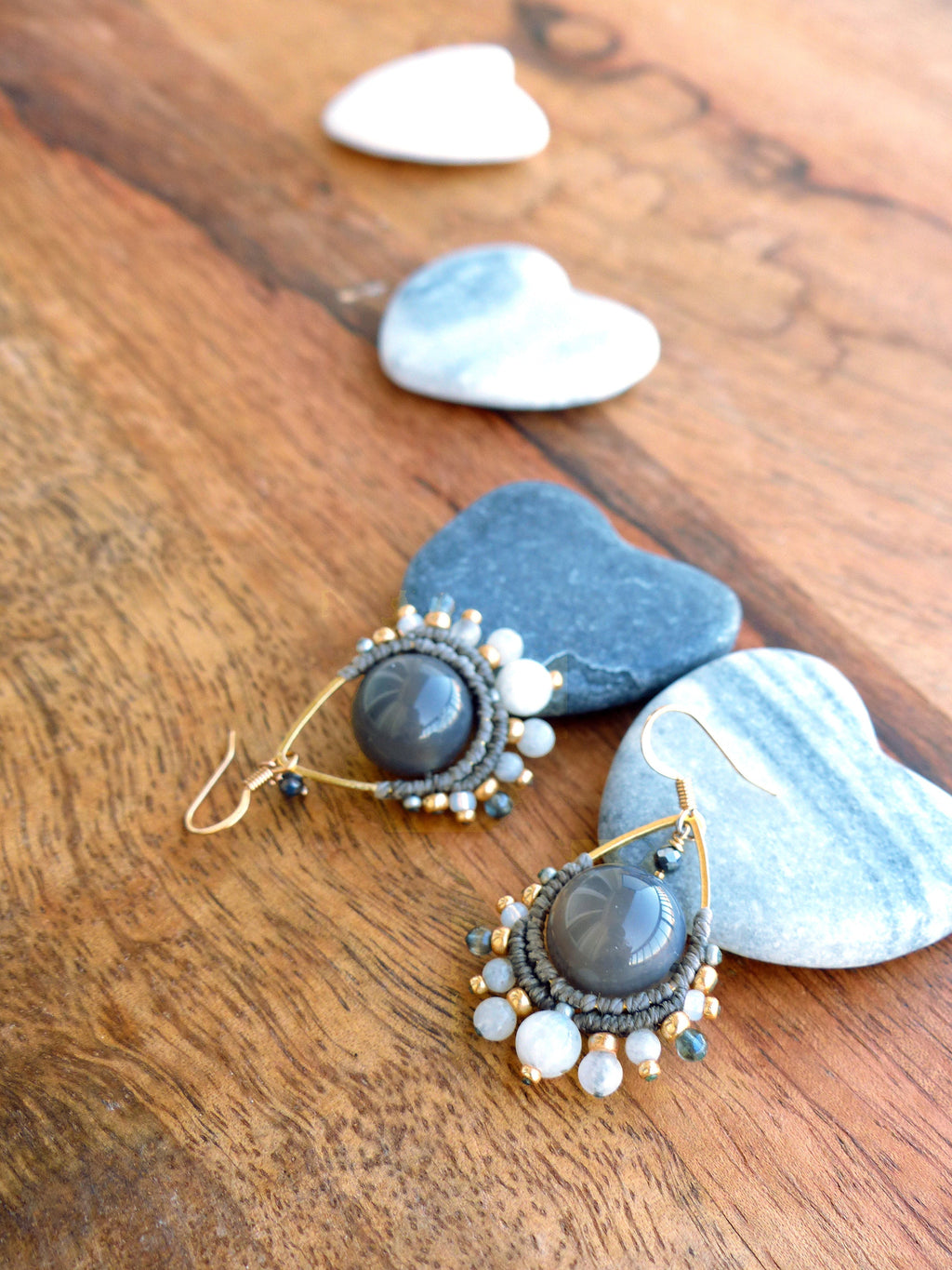 Peut inclure: Une paire de boucles d'oreilles faites à la main avec un centre en pierre grise, entouré d'un design macramé. Les boucles d'oreilles ont des crochets dorés et sont ornées de petites perles blanches et dorées. Elles sont présentées sur une surface en bois avec des pierres en forme de cœur.
