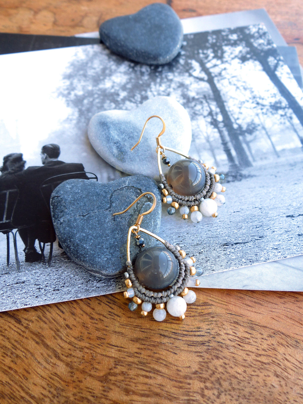 Peut inclure: Boucles d'oreilles faites à la main avec un centre en agate grise, entourées d'un design macramé. Les boucles d'oreilles comportent des crochets dorés et sont agrémentées de petites perles blanches et dorées. Les boucles d'oreilles sont exposées sur une surface en bois.