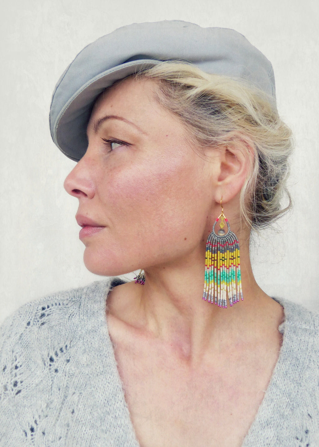 Peut inclure: Une femme portant un pull en tricot gris et une casquette plate grise. Elle porte de longues boucles d'oreilles en perles colorées en forme de goutte.