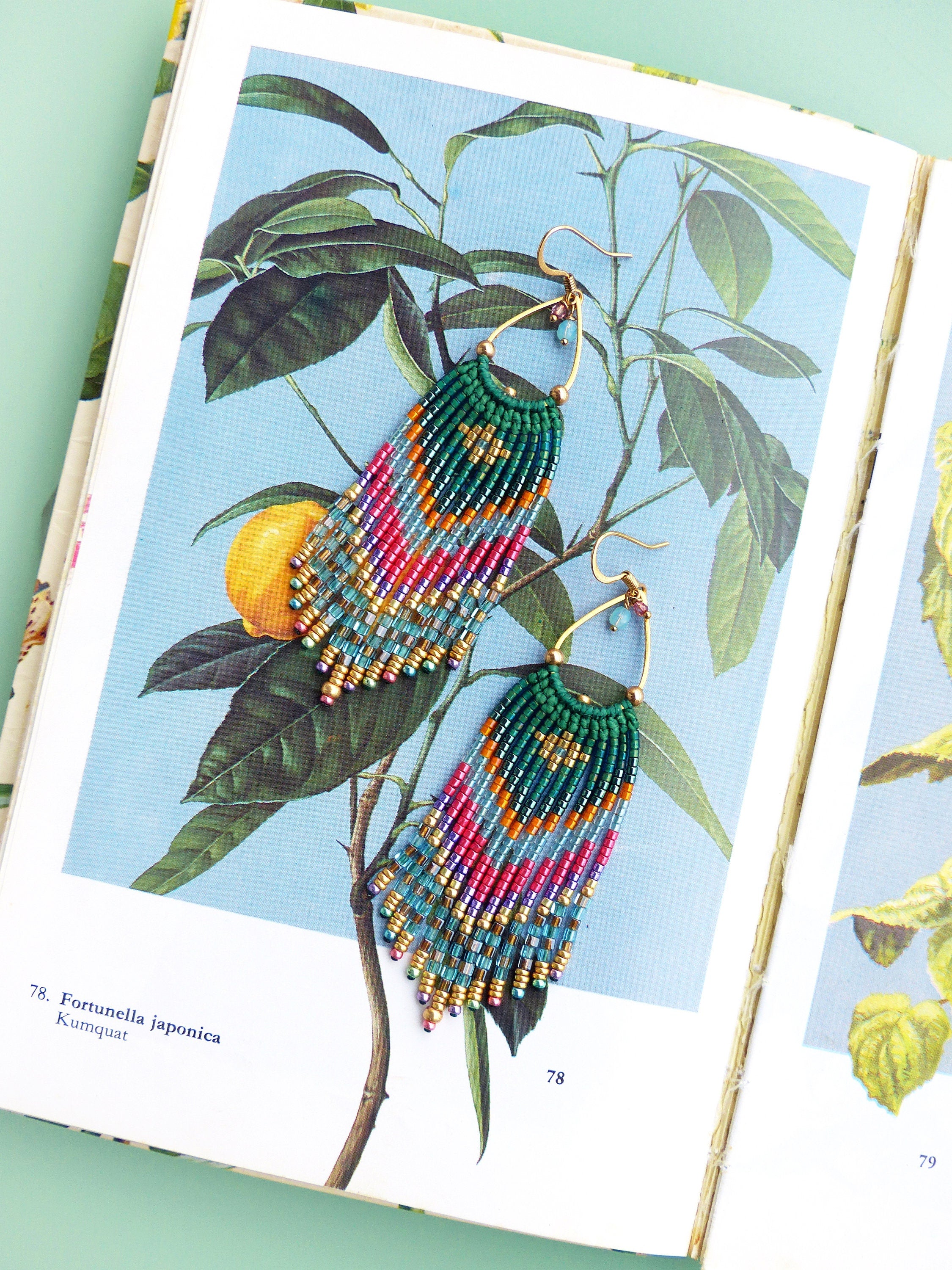 Peut inclure: Une paire de longues boucles d'oreilles en perles avec un anneau en or et une frange de perles turquoise, vertes, roses et dorées. Les boucles d'oreilles sont photographiées sur une illustration botanique d'un kumquat.
