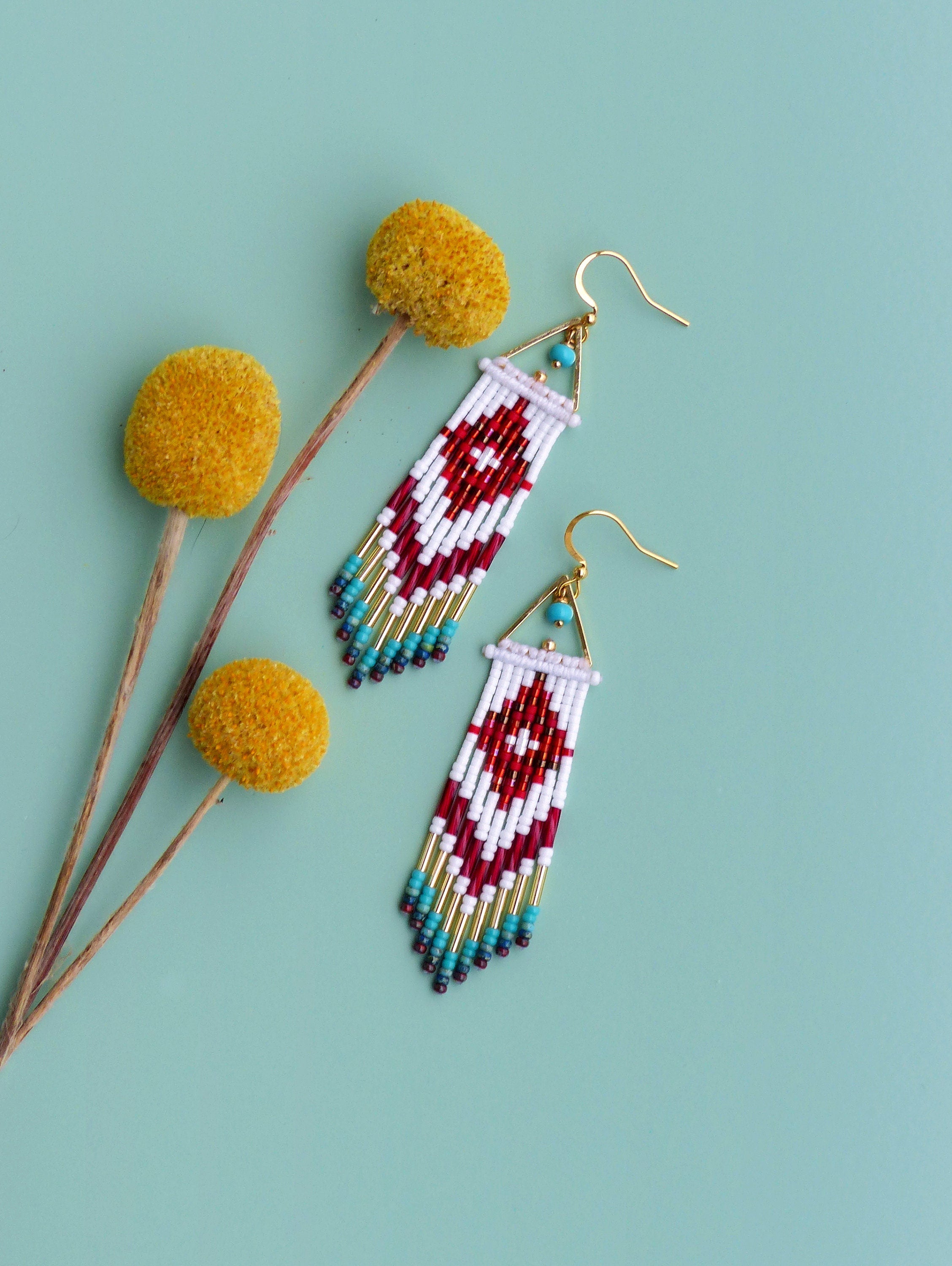 Peut inclure: Une paire de longues boucles d'oreilles en perles avec un motif géométrique en rouge, blanc et or. Les boucles d'oreilles ont une frange de perles turquoise et dorées.